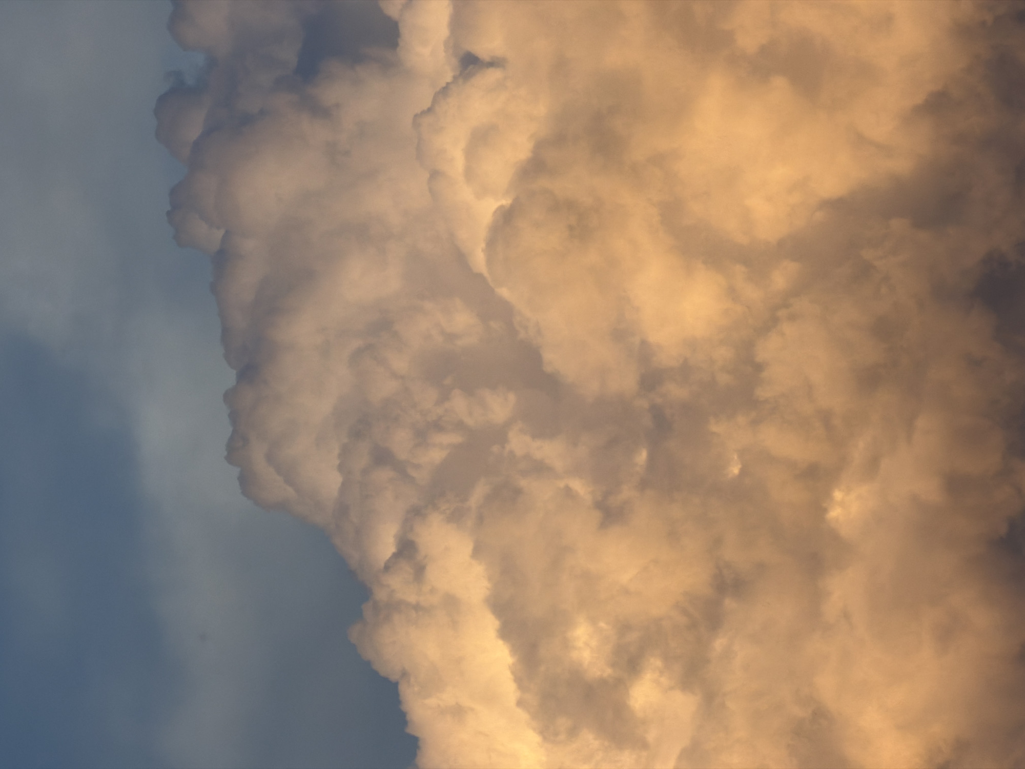 The image shows a close-up view of dramatic, illuminated clouds against a dark, blue sky. The clouds are predominantly in shades of golden orange and white, suggesting they are being lit by the sun from a low angle, possibly during sunrise or sunset. There are no people, objects, or discernible structures visible, indicating this is a natural landscape shot focusing on atmospheric conditions. The scene is devoid of any specific urban or geographical markers that would tie it to Tsiolkovsky, Russia, or any other city. The focus is entirely on the texture, form, and color of the clouds.