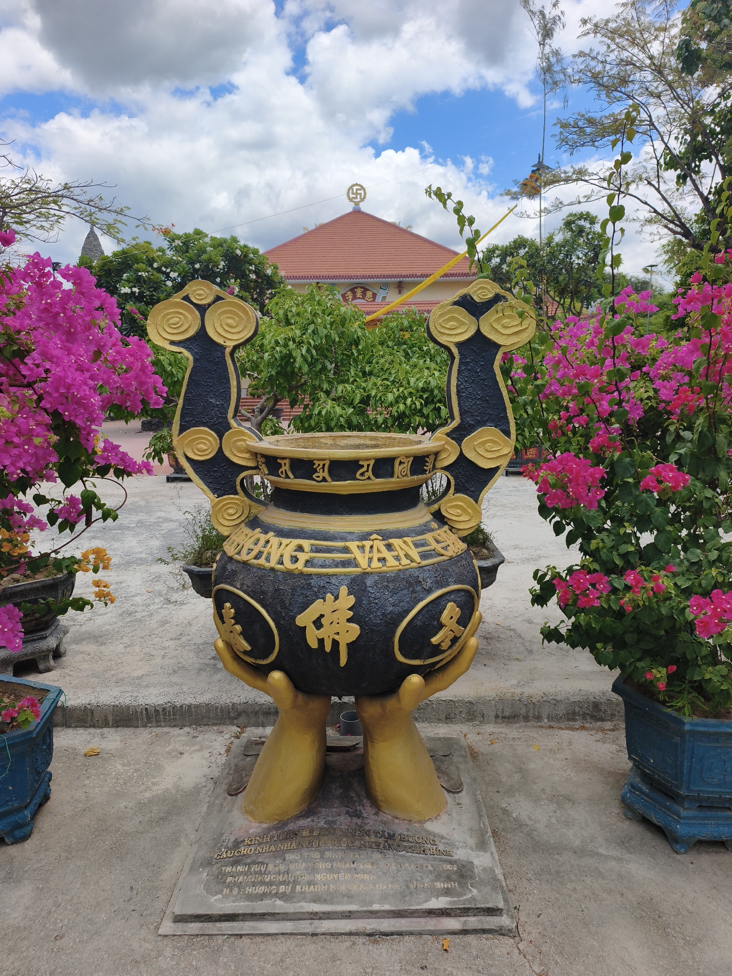 The image depicts a large, ornate incense burner supported by two golden hands. The burner is black with gold decorative elements and inscriptions, including what appears to be a Buddhist symbol and text in Vietnamese. It is situated outdoors on a paved area. In the background, there is a building with a reddish-orange tiled roof, topped with a golden Buddhist symbol. Lush green trees and vibrant pink bougainvillea bushes surround the incense burner, with one bush prominently displayed on the right side of the frame. The sky above is blue with scattered white clouds. A stone plaque with text is visible at the base of the incense burner. This scene is located at Hue Nghiem Pagoda in Cam Ranh, Khanh Hoa Province, Vietnam.