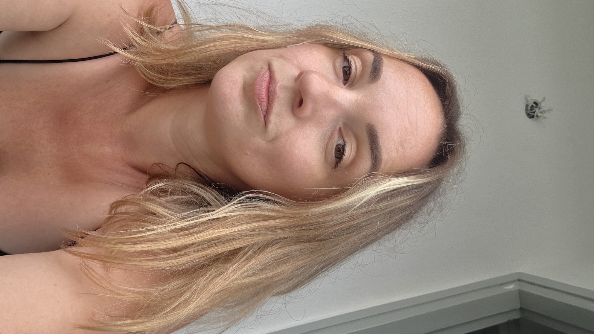 A medium shot captures a woman with blonde hair and brown eyes looking at the camera. She is wearing a black spaghetti strap top. Her skin is lightly tanned and has a few freckles. The background is a white wall with a light fixture at the top. The lighting appears to be natural. The image was taken outdoors, possibly on a balcony, given the presence of a railing visible on the right side of the frame.