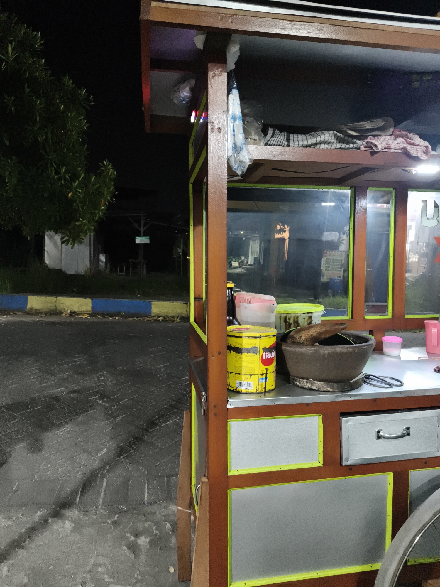 An outdoor, nighttime scene features a brightly lit street food cart, likely in Sidorejo, Indonesia. The cart, constructed from dark brown wood with bright yellow/neon green trim outlining grey/silver panels, is positioned on the right side of the frame. A bicycle wheel is partially visible at the bottom right.
The top section of the cart holds various items, including neatly folded checkered and red-patterned cloths, and a white plastic bag. Two small LED lights, one red and one blue, are affixed to the front, casting light onto the upper shelf. The middle section consists of glass panels, reflecting ambient lights and indistinct background elements.
The main countertop, made of a metallic, reflective material, displays several items for food preparation. These include a large, weathered yellow tin with partial text "Tiga" and "LIQUI..." (likely "LIQUID") visible on its label. Next to it is a traditional brown stone mortar and pestle (cobek and ulekan), suggesting the preparation of ground ingredients like spices or sauces. Other items include a dark plastic bottle, a clear plastic jug, a green plastic bucket, a pink plastic jug, and a small pink container with a white lid. Metal tongs are also present on the counter. Below the counter, the cart features a silver drawer with a handle.
The background is dark, indicating night. To the left, lush dark green foliage of a tree stands against the black sky. The ground in the foreground is a paved surface, possibly wet or reflecting light, with distinct brick-like patterns. A curb painted in blue and yellow stripes runs along the edge of the paved area. Further in the distance, obscured by darkness, are faint structures, including what appears to be a white tent-like object and a small sign on a pole, the text of which is unreadable. No people are visible in the image. The artificial lighting of the cart strongly contrasts with the surrounding darkness, highlighting its function as a nocturnal vendor.