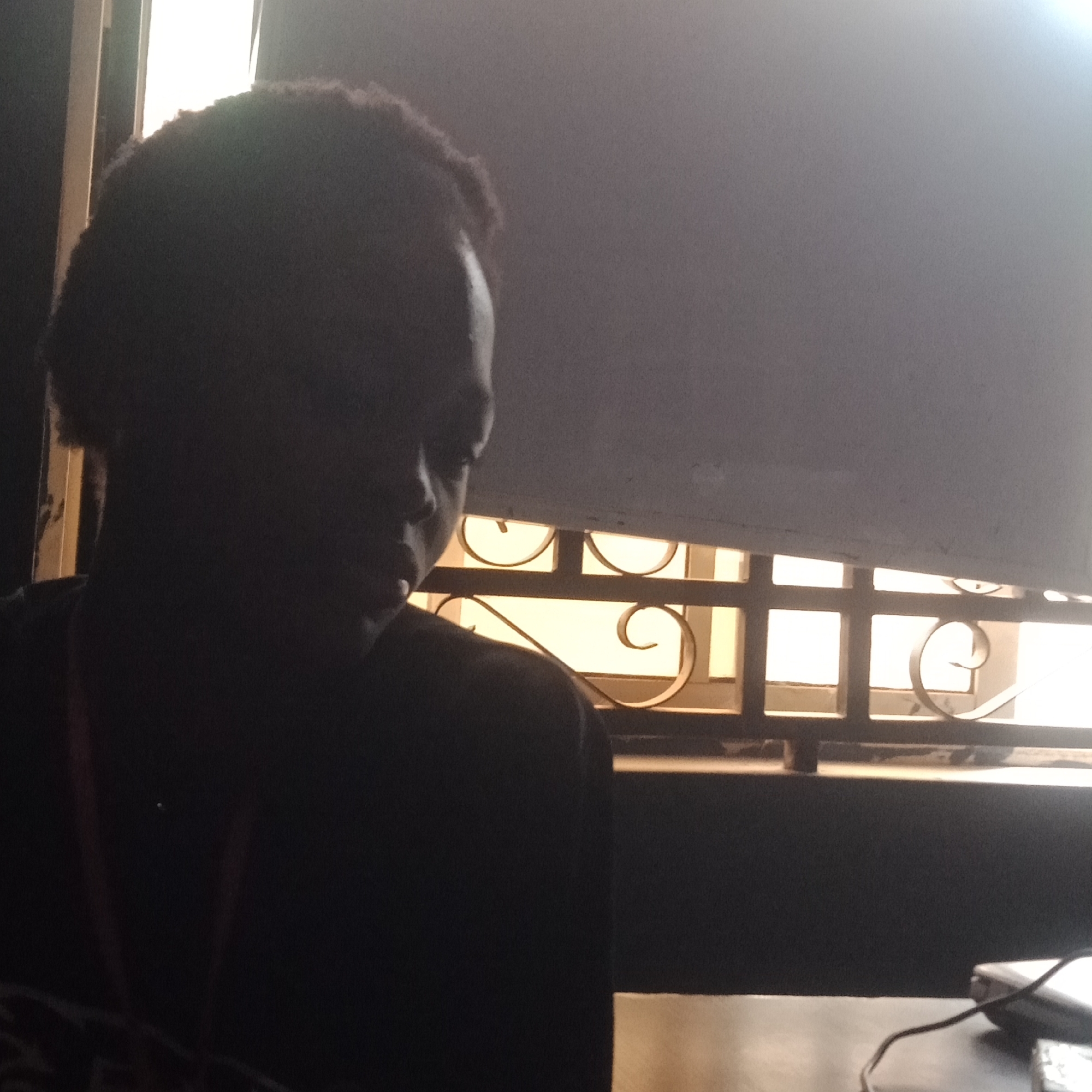 The image depicts a close-up, silhouetted view of a person from the shoulders up, facing left. The person has dark, curly hair. They are positioned indoors, with a window featuring decorative metalwork visible in the background, illuminated by daylight filtering through. The overall lighting suggests it is either early morning or late afternoon. The scene is dark, with the subject's features largely obscured by shadow. A portion of a white blind or curtain is also visible. The setting is likely a residential or office space in Mowe, Nigeria.