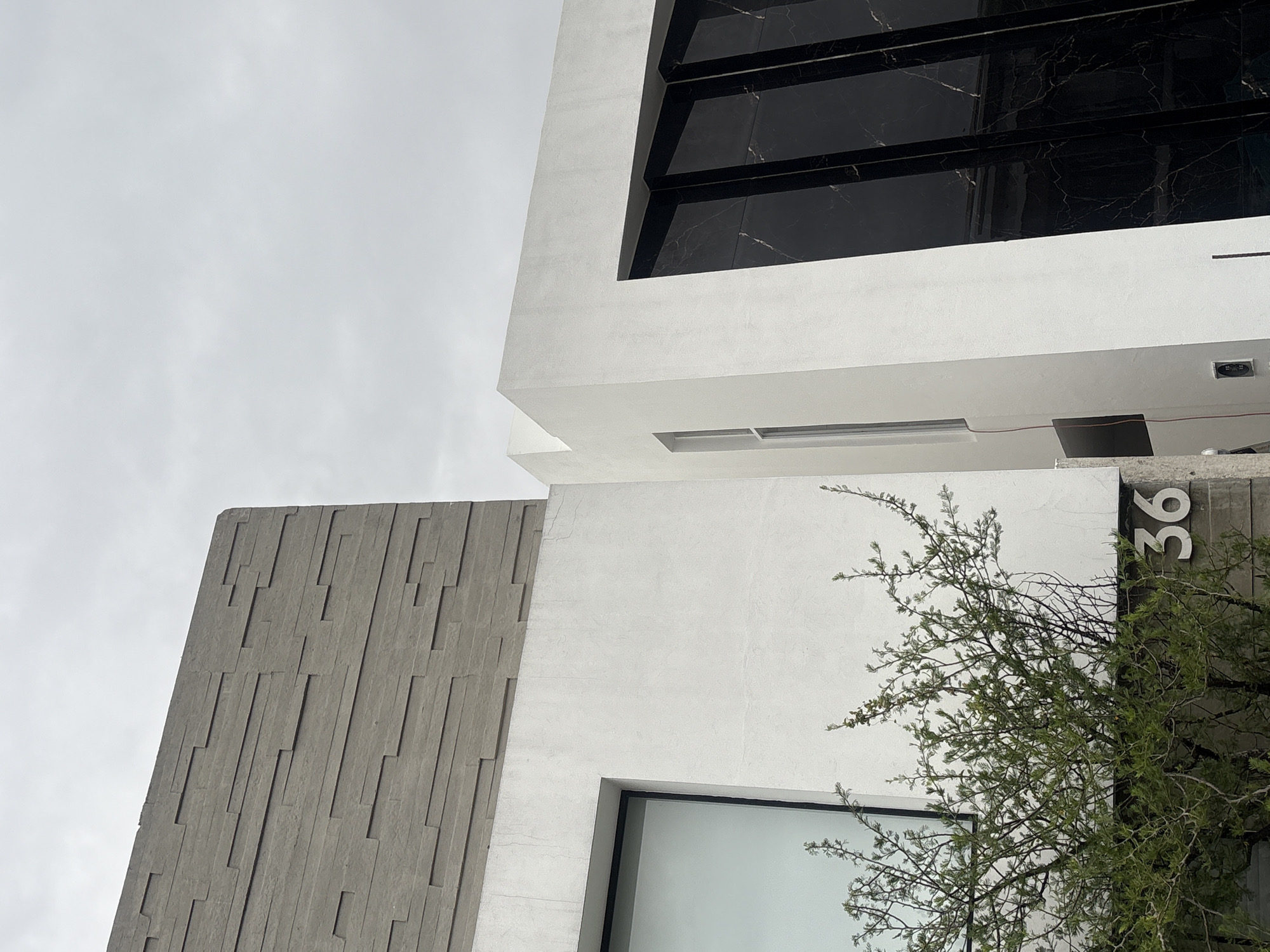 An eye-level shot of a modern building facade in San Bernardino Tlaxcalancingo, Mexico. The building features a combination of materials including a grey stone-like texture with vertical striations, smooth white walls, and black marble-like panels around a window. The address number "36" is visible on the right side of the building, partially obscured by a leafy green bush. The sky is overcast and pale grey. A window is seen on the bottom of the image.