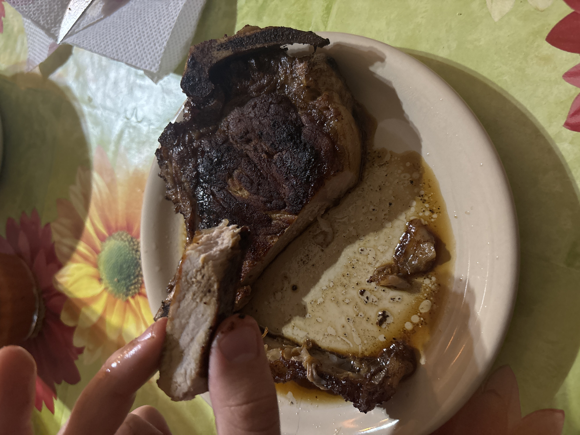 The image shows a plate with a large, cooked chop of meat, possibly pork or beef, which has been partially cut. A hand is holding a slice of the meat. There is evidence of sauce or grease on the plate. A napkin is visible in the upper left corner. The plate sits on a patterned tablecloth with floral designs. I am unable to determine from the image whether the location is Goritsa, Bulgaria.
