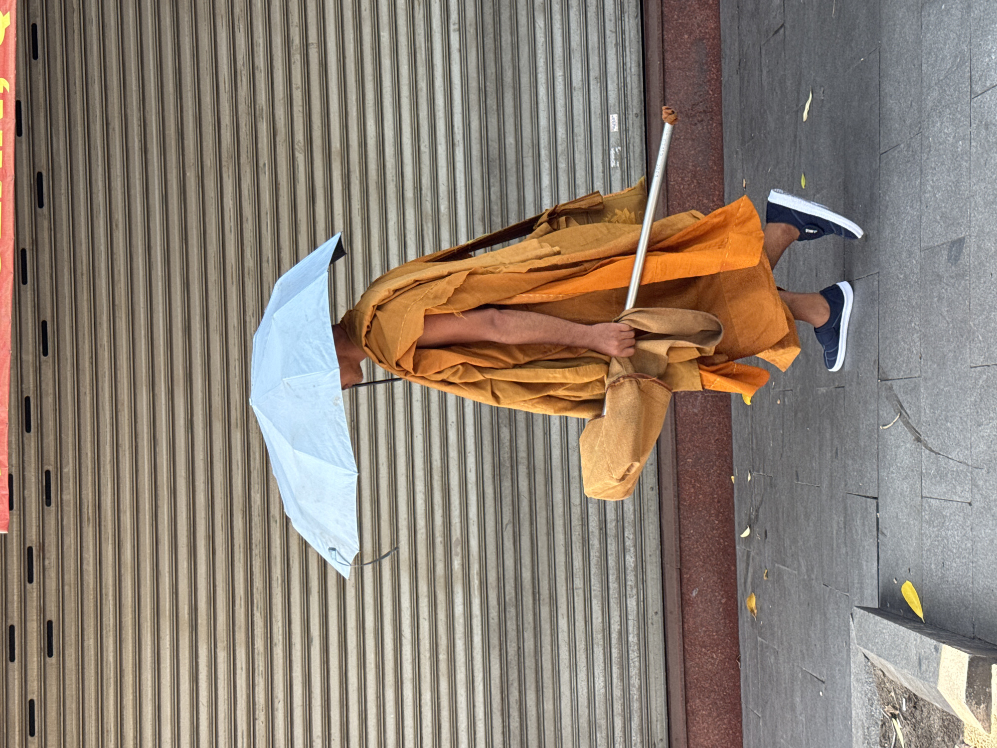 A Buddhist monk wearing an orange robe and carrying a light blue umbrella walks along a tiled sidewalk in front of a closed metal security shutter. The monk is also carrying a brown bundle under one arm and a metal pole in his right hand. The scene appears to be in Nha Trang, Vietnam, given the presence of the security shutter style common in the region and the warm, sunny climate suggested by the umbrella use. There is some litter on the sidewalk, including yellow leaves. The overall impression is one of a street scene, possibly during a midday walk or procession.
