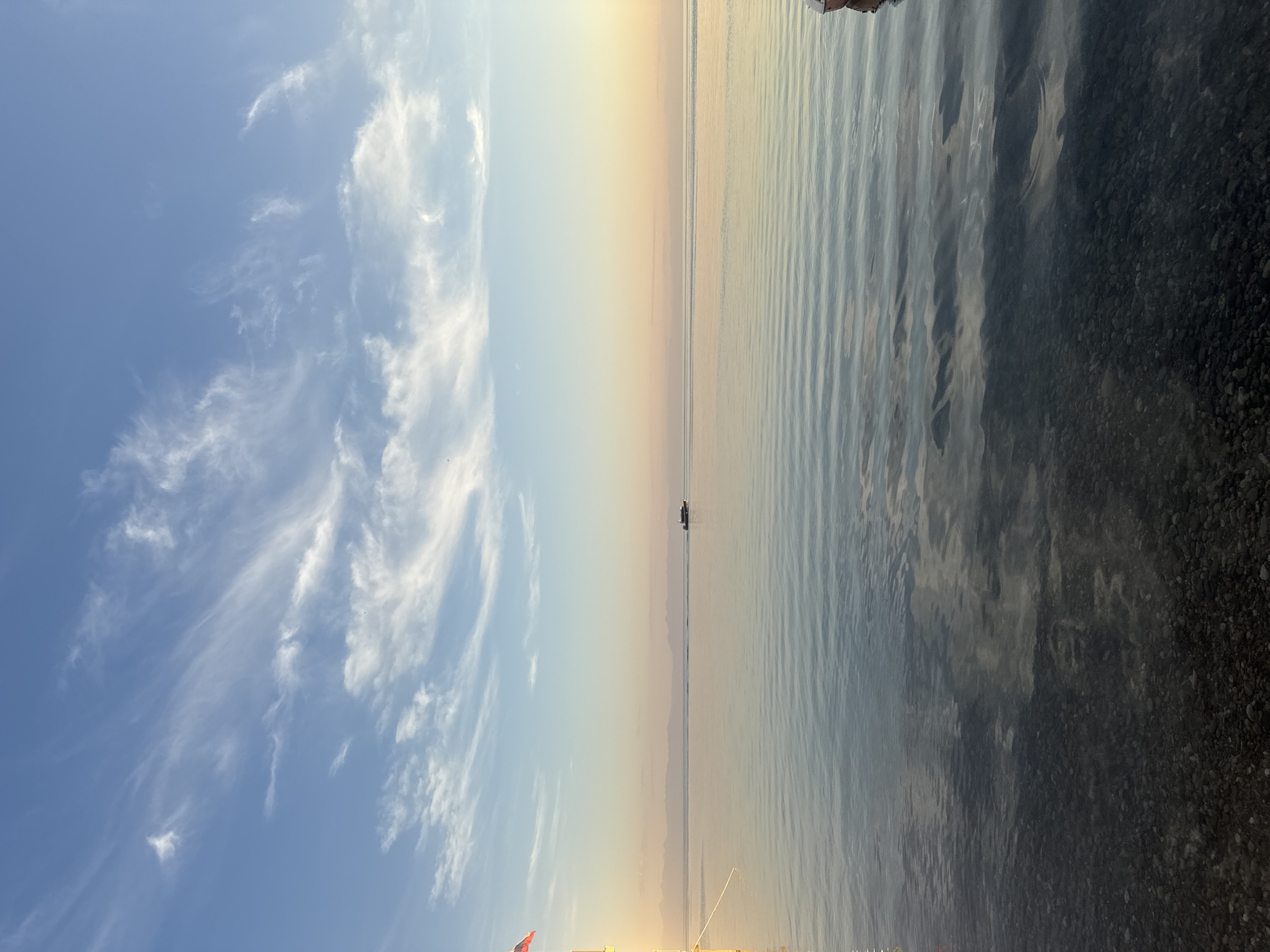 The image depicts a tranquil scene at Lake Baikal in Listvyanka, Russia, likely during sunrise or sunset. The sky is a gradient of pale blue near the top, transitioning to soft oranges and yellows closer to the horizon. Wispy white clouds are scattered across the upper portion of the sky, catching the warm light.
The horizon is marked by a thin line of distant mountains, appearing as a muted silhouette against the colorful sky. A small motorboat is visible on the calm, expansive water, moving horizontally across the frame in the middle distance.
In the foreground on the left, a portion of a yellow structure, possibly a dock or a building, is visible, with a rope extending from it and a flag with red, white, and blue stripes (likely the Russian flag) attached. The water in the foreground is clear, revealing a bed of dark, smooth pebbles beneath its surface. The surface of the lake is almost perfectly still, reflecting the sky and distant mountains with subtle ripples. On the far right edge of the image, a small portion of a red and white object, possibly another boat or buoy, is visible. The overall atmosphere is one of peace and natural beauty.