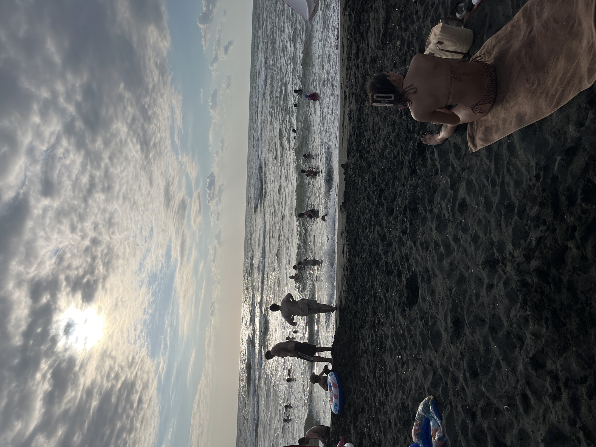 The image depicts a crowded beach scene in Ureki, Georgia, known for its magnetic sand. The sun is partially obscured by clouds, casting a soft light over the water and sky. Several people are visible in the ocean, swimming and playing in the waves. On the dark sand beach, individuals are seen relaxing, with one person in the foreground sitting on a towel, facing away from the camera, wearing a bikini. In the mid-ground, a group of people, including adults and children, are standing near the water's edge, some holding inflatable toys. The overall atmosphere suggests a lively summer day at the beach.