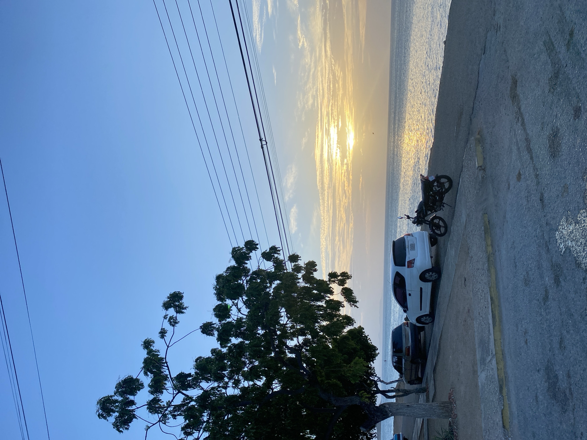 The image depicts a coastal scene in Juan Griego, Venezuela, during what appears to be either sunrise or sunset. The sun's light reflects on the water surface. Several power lines stretch across the sky. A large tree dominates the left foreground. Adjacent to the water, parked on the side of the road, are a motorcycle, a white car, and a dark-colored vehicle. The road surface is concrete.
