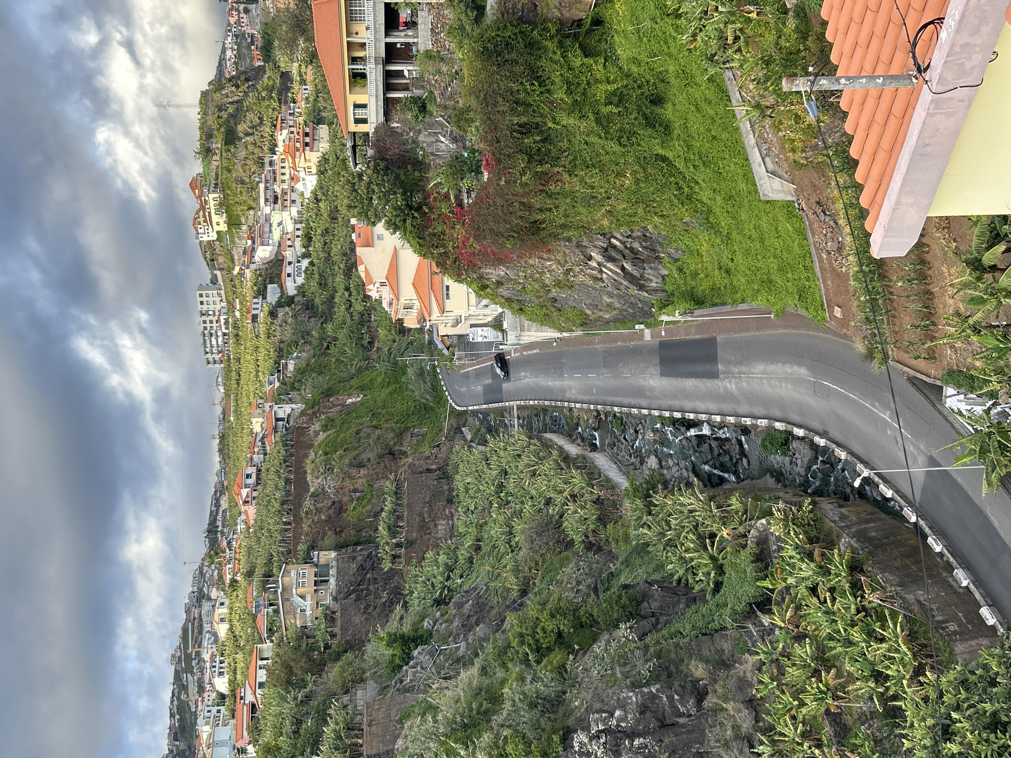 This image captures a view of the coastal town of Câmara de Lobos, Portugal. The scene is characterized by a winding road curving along the side of a steep, rocky hillside. Buildings with terracotta roofs are interspersed along the slope, nestled among dense green vegetation. Below the road, a small waterfall or stream cascades down the rocks. In the distance, additional buildings extend up the hillside under a cloudy sky. A single vehicle can be seen on the road. The overall impression is a landscape where urban development meets natural terrain.