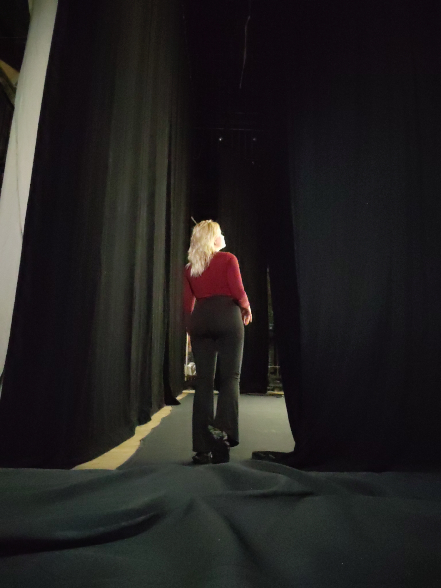 The image depicts a backstage or performance area, likely within a theater or event hall in Izhevsk, Russia. A person, appearing to be a woman with blonde hair, is captured from behind, standing between parted heavy black curtains. She is dressed in a dark red long-sleeved top and dark, possibly black, trousers. Her gaze is directed upwards and forward into an unseen, brightly lit space, suggesting she is observing a performance, preparing to go on stage, or looking into an adjacent area.
The setting is characterized by tall, dark black curtains that flank both sides, creating a narrow corridor or pathway. On the far left, a vertical strip of a lighter-colored, possibly white or cream, curtain or panel is visible. The floor is covered with dark fabric, showing some folds and variations in texture, especially in the foreground. The lighting in the immediate area around the person is dim, casting a subdued atmosphere that contrasts with the brighter light originating from beyond the parting curtains. This contrast emphasizes the transition from the dark backstage to a more illuminated front area. The overall scene conveys a sense of anticipation or quiet focus typically found behind the scenes of a performance. No text is visible in the image.