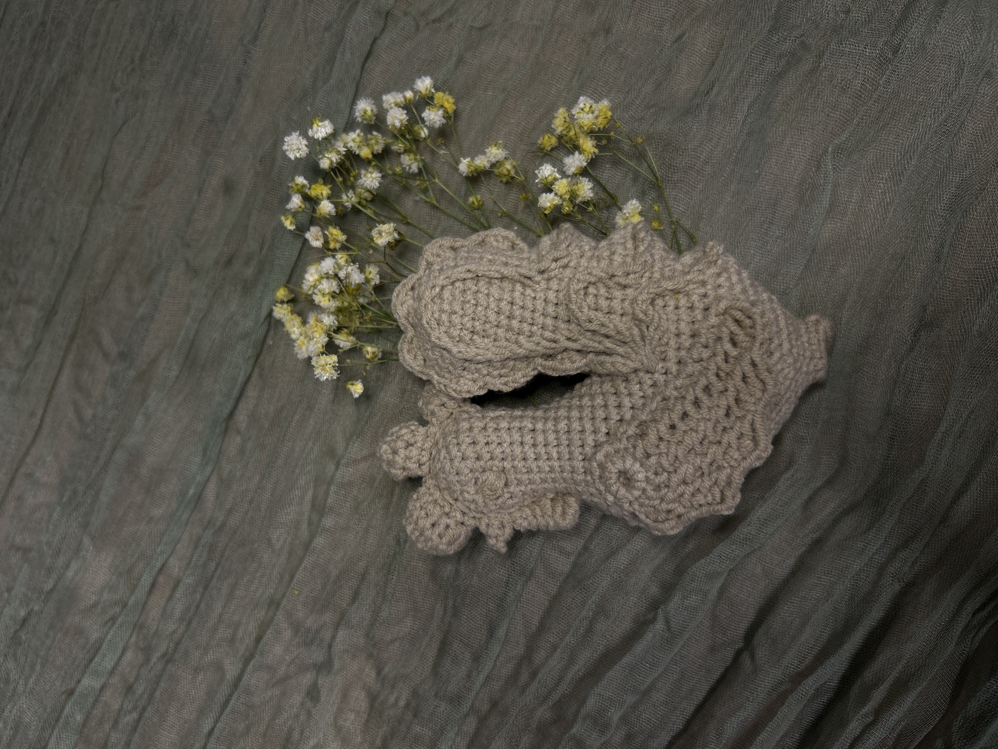 This still life image, captured in Saint Petersburg, Russia, features two light brown or beige crocheted items and a small cluster of dried white and yellow flowers, all arranged on a dark gray-green, crinkled sheer fabric background.
The main subjects are the two handcrafted crocheted pieces, which appear to be baby booties or small soft footwear. They are made from yarn with a visible, textured crochet stitch pattern. One piece is positioned slightly behind and to the left of the other. Both show scalloped or decorative edges, giving them a delicate appearance.
Interspersed with the crocheted items, primarily emerging from behind the upper one, is a sprig of dried flowers resembling Baby's Breath (Gypsophila). These flowers are small and numerous, with both white and faded yellow blooms, adding a touch of natural beauty and softness to the composition.
The background is a dark, muted gray-green fabric with a sheer, crinkled texture, possibly gauze or muslin. Its folds and ripples create subtle shadows and add depth to the scene.
The image depicts a static arrangement, likely intended for product photography or as a decorative display. The overall tone is calm and gentle, highlighting the texture of the crocheted items and the delicate nature of the flowers against the soft, diffused background. There is no visible text in the image. The lighting appears soft and even, suggesting an indoor setting with no specific time of day or weather conditions discernible.