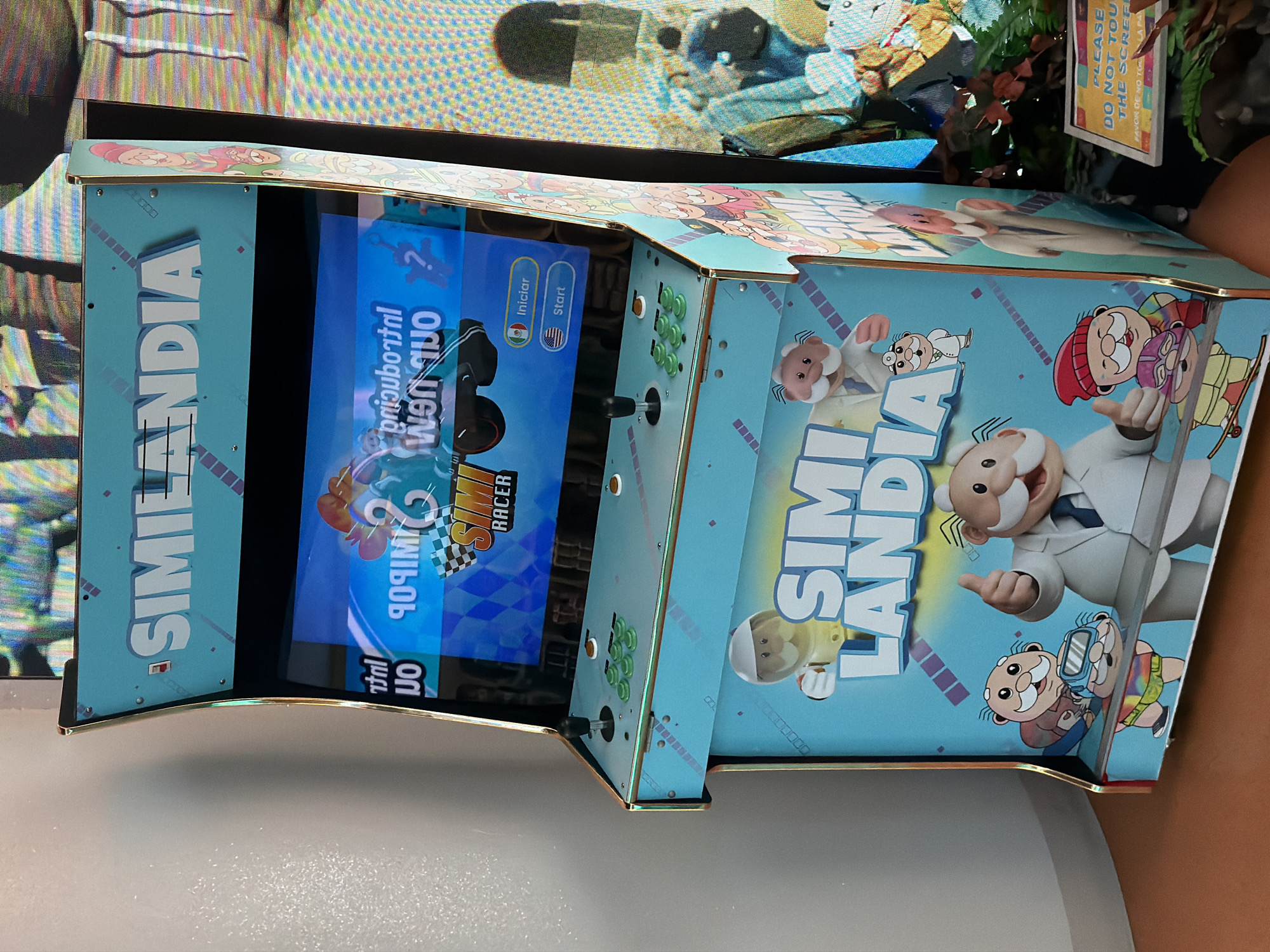 The image depicts a light blue arcade game machine labeled "Similandia" with cartoon characters. The screen displays the game "Simi Racer," with options to choose between Spanish and English languages for the start menu. The game console has a joystick and green buttons. A sign that reads, "Please do not touch the screen." It is placed next to a potted plant. The background is blurry, but the scene may be in a game room or arcade, possibly in Los Angeles.