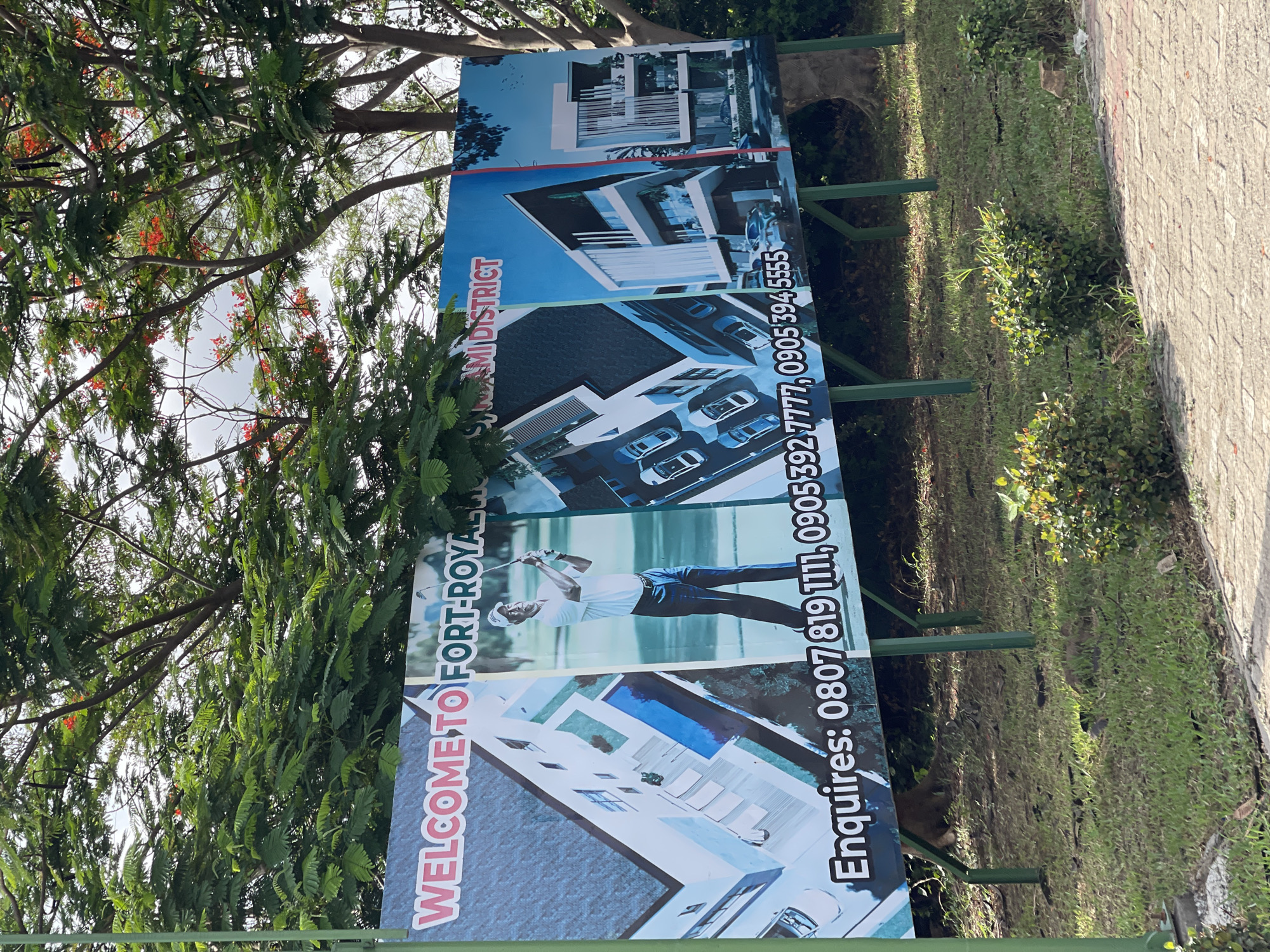 The image depicts a rectangular advertising sign installed on green metal poles. The sign is positioned on a grassy area beside a paved walkway in Gosa, Nigeria. The sign displays the words "WELCOME TO FORT-ROYAL NAMI DISTRICT" in red, white and blue lettering, followed by images of modern buildings and a golfer, indicating real estate development. Contact information "Enquiries: 0807 819 1111, 0905392 7777, 0905394 5555" is displayed. Foliage from a large tree obscures part of the sign and sky above.