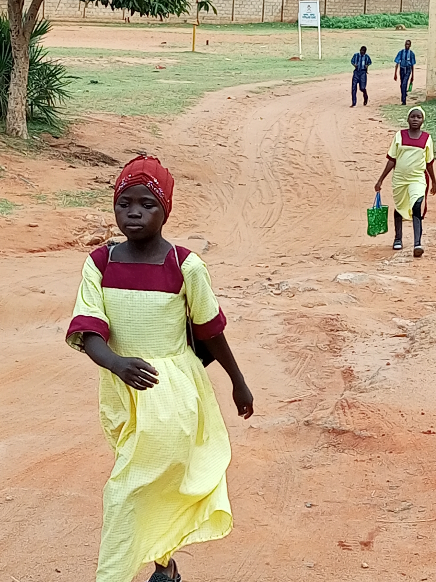 An outdoor, eye-level, medium shot captures four individuals on a dusty, unpaved road in Gombe, Nigeria. The foreground is dominated by a young girl, facing left and slightly away from the camera. She wears a light yellow dress with maroon trim on the shoulders and sleeves, and a matching maroon headscarf. Her expression is somber. Behind her and to the right, another girl, also wearing a light yellow dress with maroon trim, walks away from the camera carrying a green patterned shopping bag. Further in the background, two young males in dark blue uniforms with light blue stripes walk away from the camera. They appear to be walking on a path with sparse green grass on either side. A pale, rectangular signpost stands on the left side of the background, and a brick wall forms a fence behind it. The road is a reddish-brown dirt with tire tracks visible. The lighting suggests it is daytime.