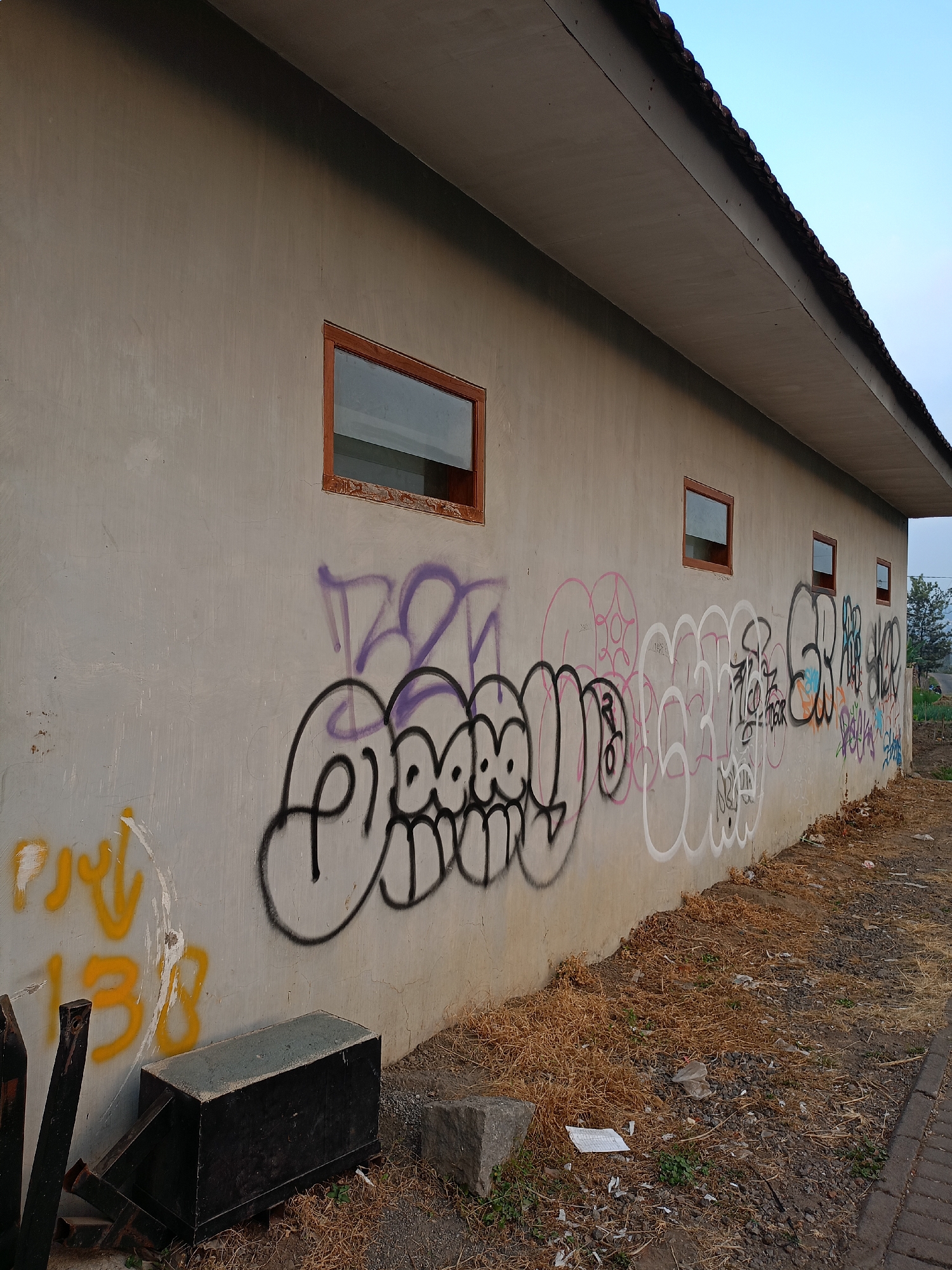 In the city of Batu, Indonesia, a plain concrete wall is heavily defaced with graffiti in various colors, including black, purple, pink, and orange. The graffiti covers a significant portion of the wall, featuring tags, stylized lettering, and abstract designs. Several rectangular windows with wooden frames are present on the wall, some reflecting the sky. The ground in front of the wall is covered with dry grass, gravel, and scattered debris, including plastic bags and paper. To the left, there's a dark, rectangular metal object, possibly a bin or storage unit, and some metal rebar. In the background, a few trees and a clear sky are visible. The overall scene depicts an urban or semi-urban exterior with signs of neglect and artistic vandalism.