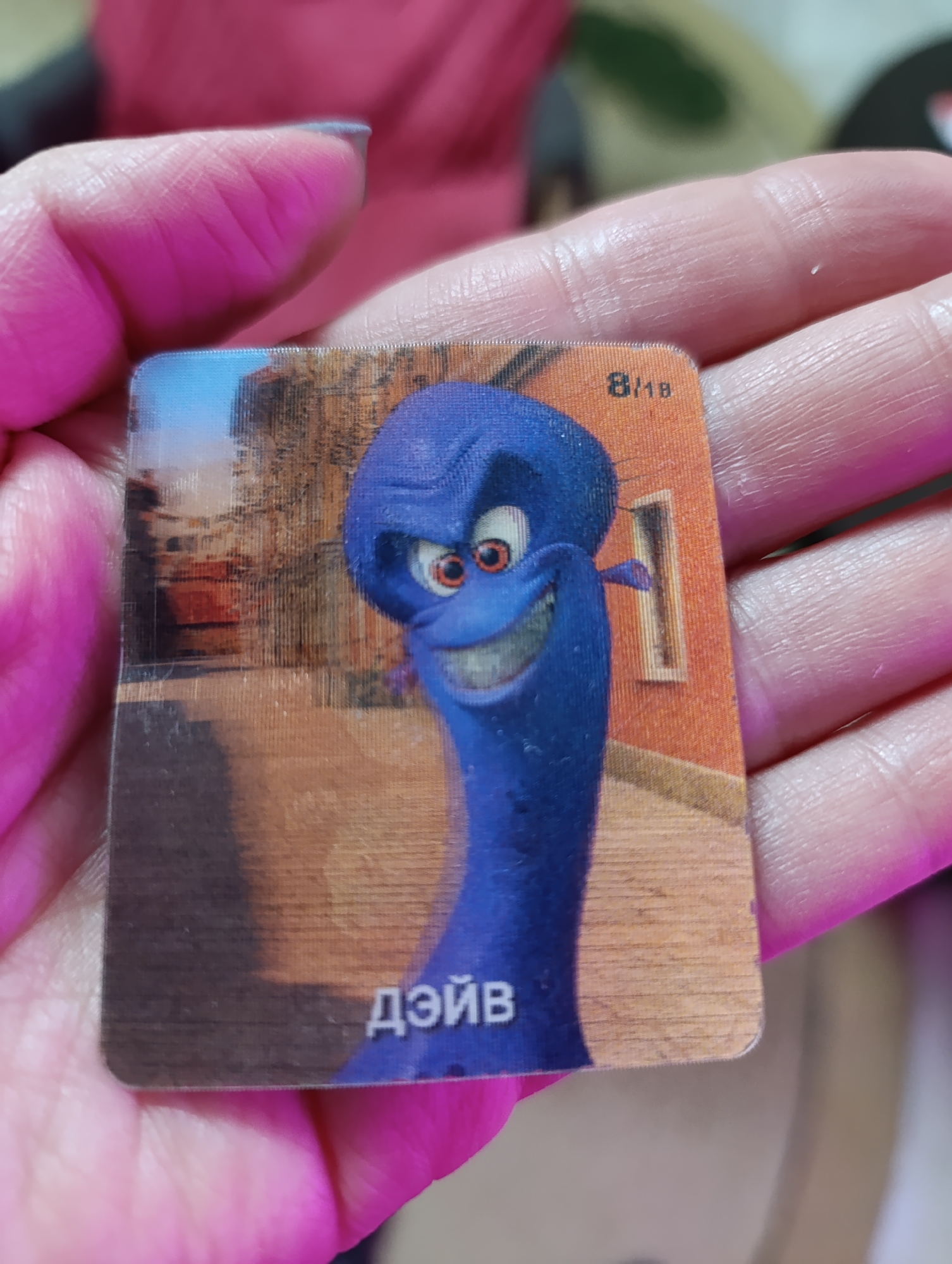 A square, laminated card featuring an animated blue character named “Дэйв” (Dave) is held in a person's hand. The card also shows the number “8/18”. The card's backdrop includes a city street with buildings painted in shades of brown and orange. The location is likely Уральский, Russia as the text is in Russian. The hand is holding the card in a well-lit area.
