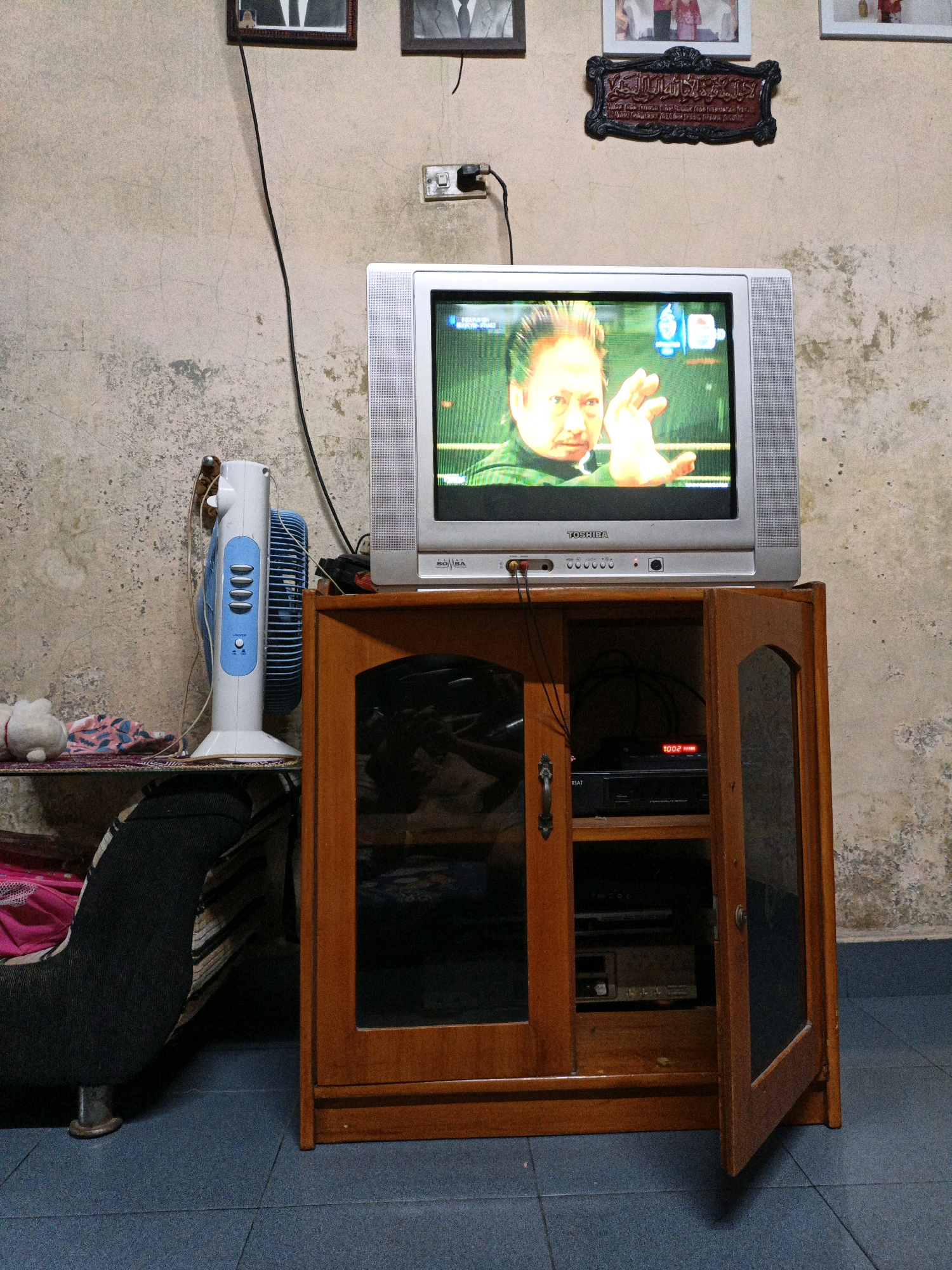 The image depicts an indoor scene in what appears to be a modest living space in Bunut, Indonesia. Dominating the center is a vintage television set, displaying a man with his hand raised, possibly from a martial arts film or program. The television rests atop a wooden cabinet with a glass-fronted door, which is partially open, revealing electronic devices inside. To the left of the television, a white and blue electric fan stands on a shelf, next to some colorful fabric and a stuffed toy. The walls are textured and aged, with visible cracks and peeling paint, and a few framed pictures hang on the wall above the television. Electrical cords snake down the wall, connecting to the television and possibly other devices. The floor is tiled in a dark blue hue. The overall impression is of a lived-in room with older electronics and decor.
