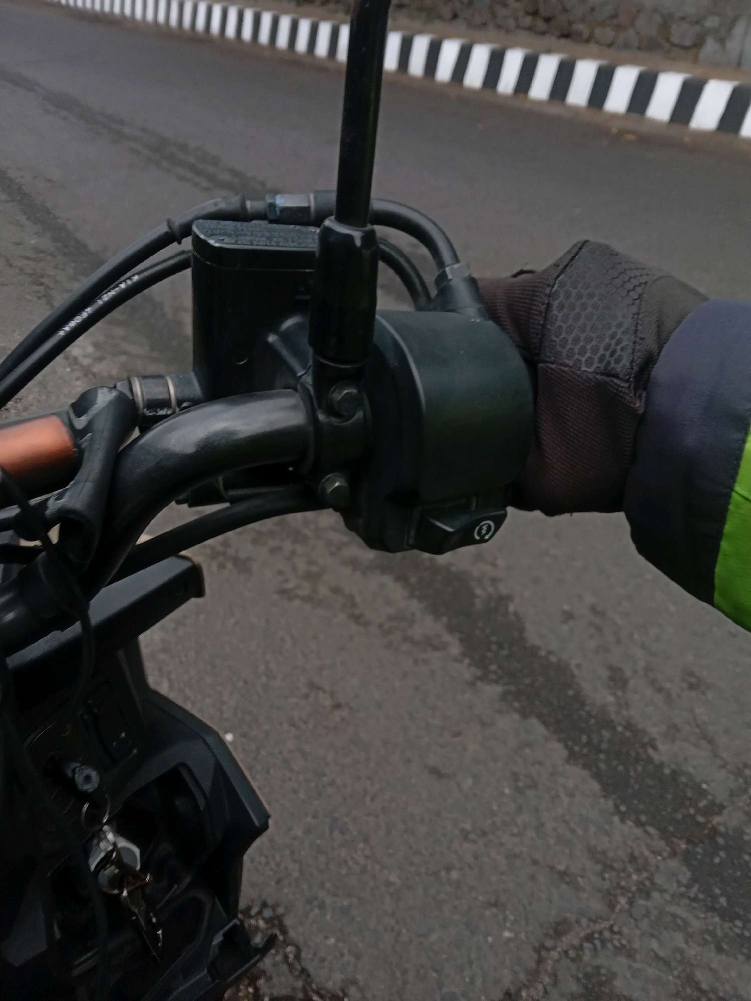 A first-person perspective shot from a motorcycle in Yogyakarta, Indonesia. The road ahead features a distinct black and white striped curb. The motorcycle's handlebar, including the brake lever and throttle, is visible. A hand wearing a black glove with hexagonal patterns grips the throttle. Part of the motorcycle's dashboard and keys are visible in the lower left corner.