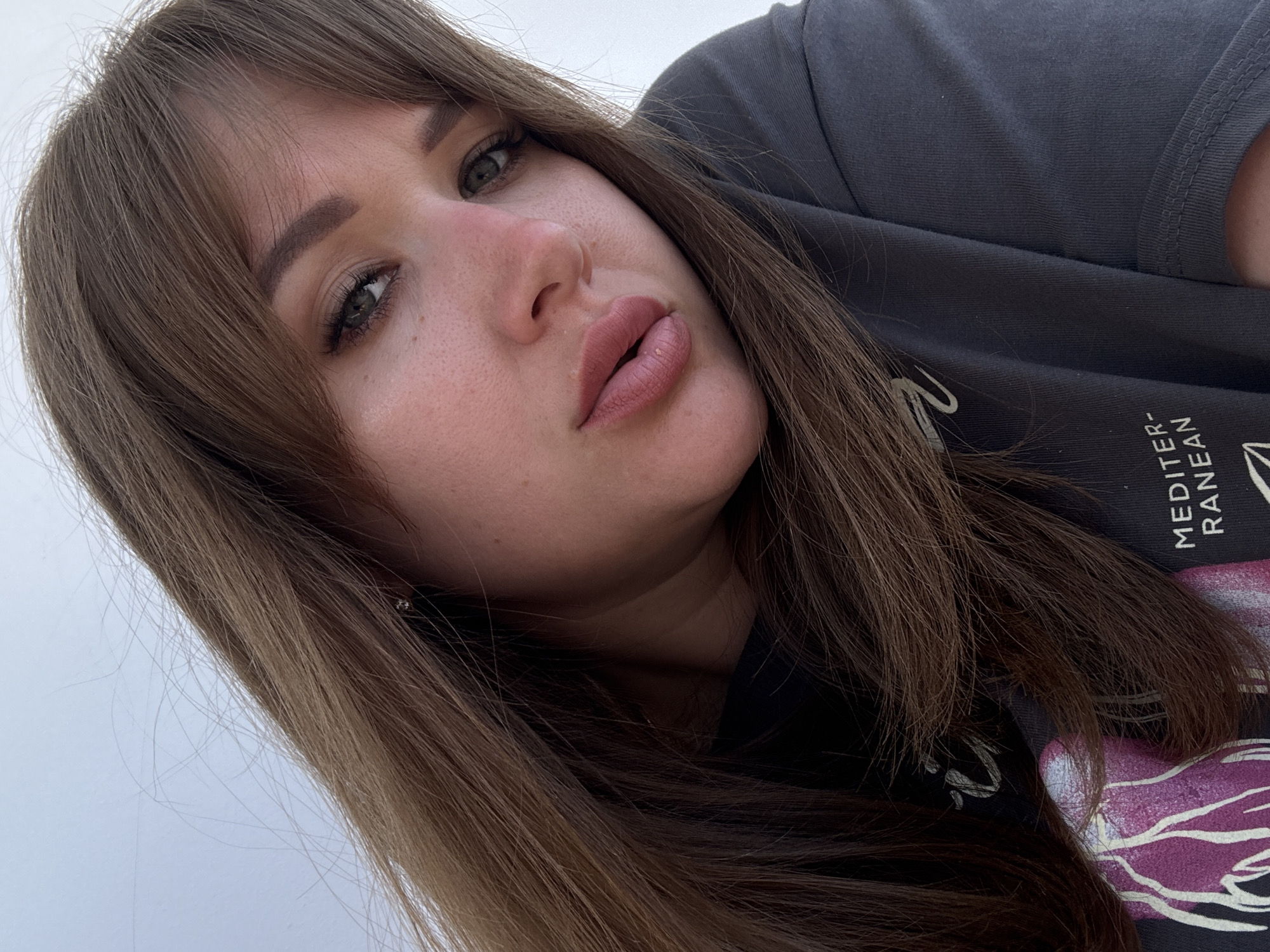 A young woman with light brown hair and green eyes is the subject of a close-up, angled selfie. She is wearing a gray t-shirt with some printed text visible. The location is indeterminate; there are no clues to suggest the city of Yevpatoriya, Ukraine. The background appears to be an overcast sky or a plain white surface.