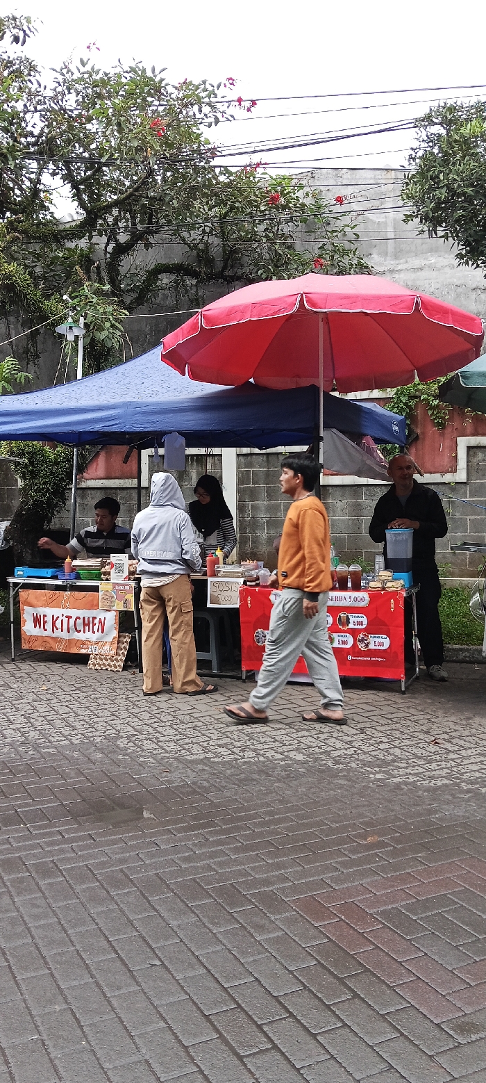 The scene is in front of a food stall in the city of Banjaran, Indonesia (latitude: -7.047, longitude: 107.58). The stall is under a blue canopy and a red umbrella. There are four people in the scene. Two are standing behind the food stall, and two are in front of it. One person is standing by a cooler, which contains a large container of ice. They are all wearing casual clothing. The stall features signs displaying the name “We Kitchen” and prices for food items. The stall is on a brick walkway. The background shows trees, a wall, and power lines. The sky is overcast.
