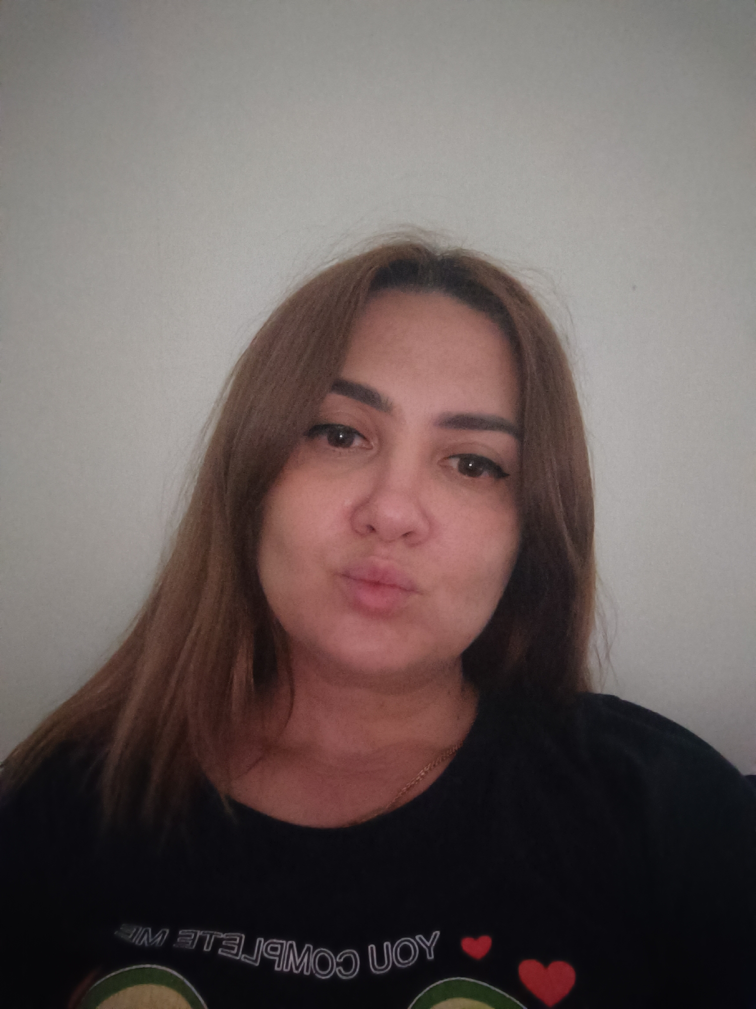 A medium shot shows a woman from the chest up, posing for a selfie against a plain, light-colored wall. She has shoulder-length, reddish-brown hair that frames her face. Her eyes are dark, and she is wearing eyeliner. Her lips are pursed. She is wearing a black t-shirt with white lettering and graphics, including red hearts and what appears to be an avocado design. The background offers no specific environmental details to place the location within Taraz city administration, Kazakhstan.