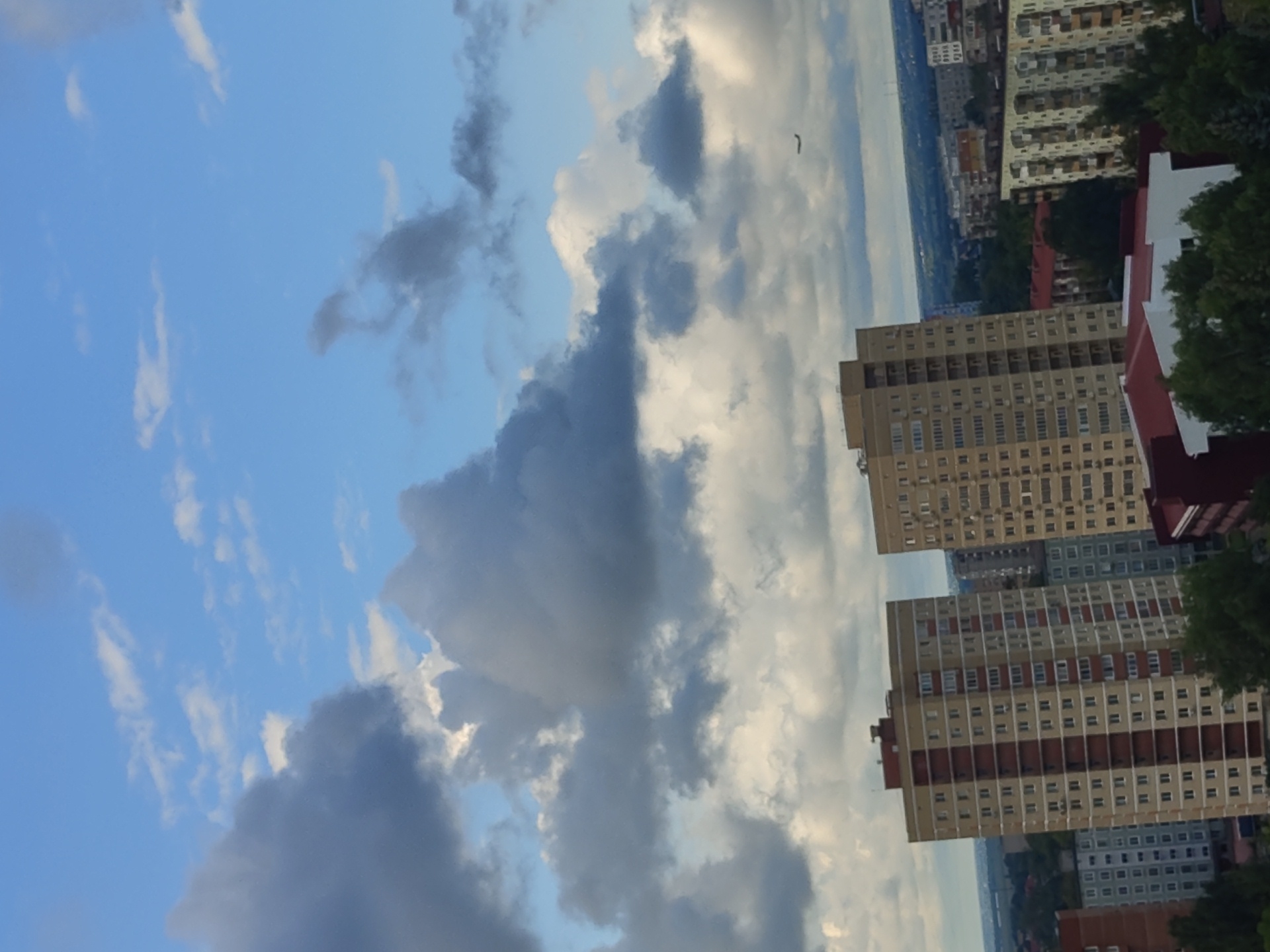 The image depicts a cityscape with numerous apartment buildings under a partially cloudy sky. In the foreground, two tall, multi-story apartment buildings are prominently featured. The building on the left is beige with vertical red stripes and numerous windows. The building on the right is also tall and beige with a more uniform window pattern. In the background, a variety of other buildings are visible, ranging in height and architectural style, interspersed with trees and a distant horizon line. A small airplane is visible in the sky on the right side of the image. The sky is a mix of blue sky and large, dark gray clouds, with some white clouds scattered throughout. The overall impression is that of a residential area in a city on a partly cloudy day. Based on the architecture, this scene is likely located in the city of Ramenskoye, Russia, given its characteristic residential building styles.