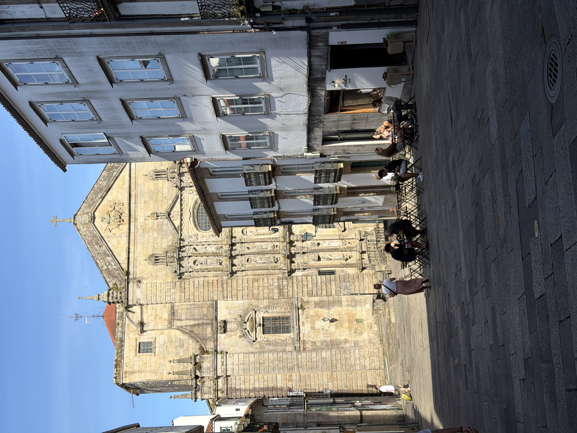 The image depicts a sun-drenched street scene in Santiago de Compostela, Spain. In the background, the imposing stone facade of a historical church, likely the Santiago Cathedral, dominates the view, with its intricate architectural details and a cross atop its roof. In the foreground, a narrow cobblestone street slopes downwards, lined with traditional European buildings. On the right, a multi-story building features a row of windows and balconies with wrought-iron railings. A cafe on the ground floor has outdoor seating with several patrons enjoying drinks and conversation. On the left, another building displays similar architectural elements. Several people are present: a person in a white shirt and shorts walks with a yellow suitcase, another person in a long skirt stands near tables, and a group of people are seated at the outdoor cafe. The sunlight casts distinct shadows across the cobblestone pavement, indicating a bright, clear day. The overall atmosphere is that of a vibrant European city square.