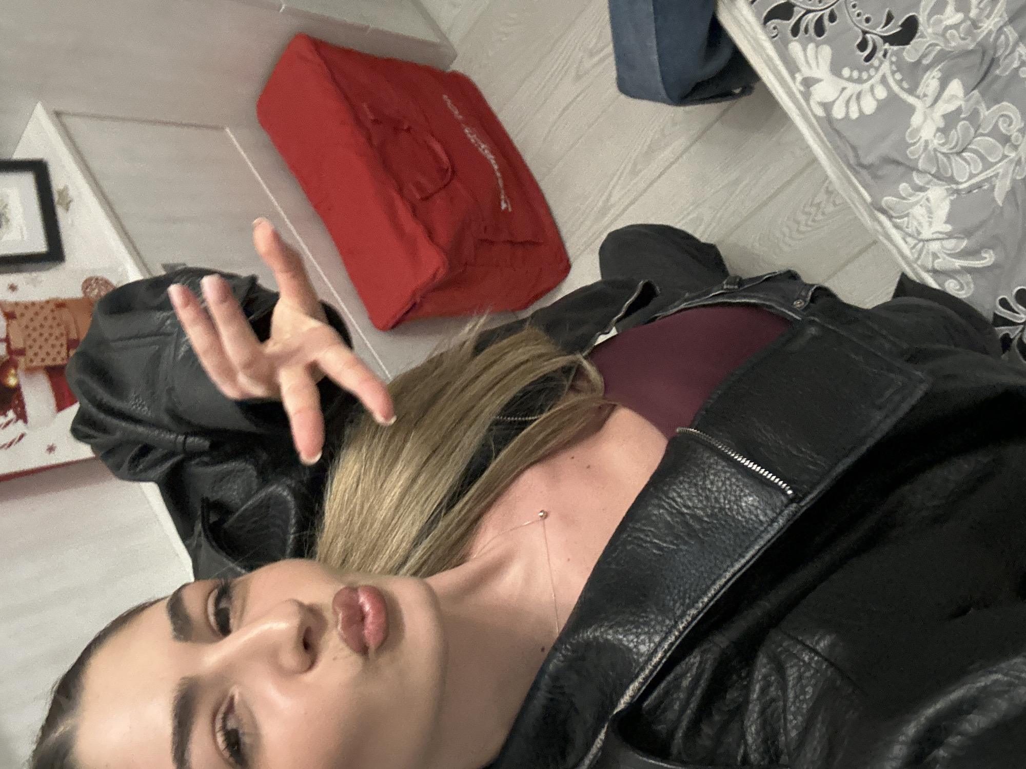 A woman lies on the floor with a black leather jacket. Her blonde hair is loose, and she wears a red top underneath the jacket. She holds up her hand with fingers spread. Next to her is a red rectangular object, possibly a cushion, on a wooden floor. A bed with a patterned comforter is visible on the right. The location is Unknown, Unknown.