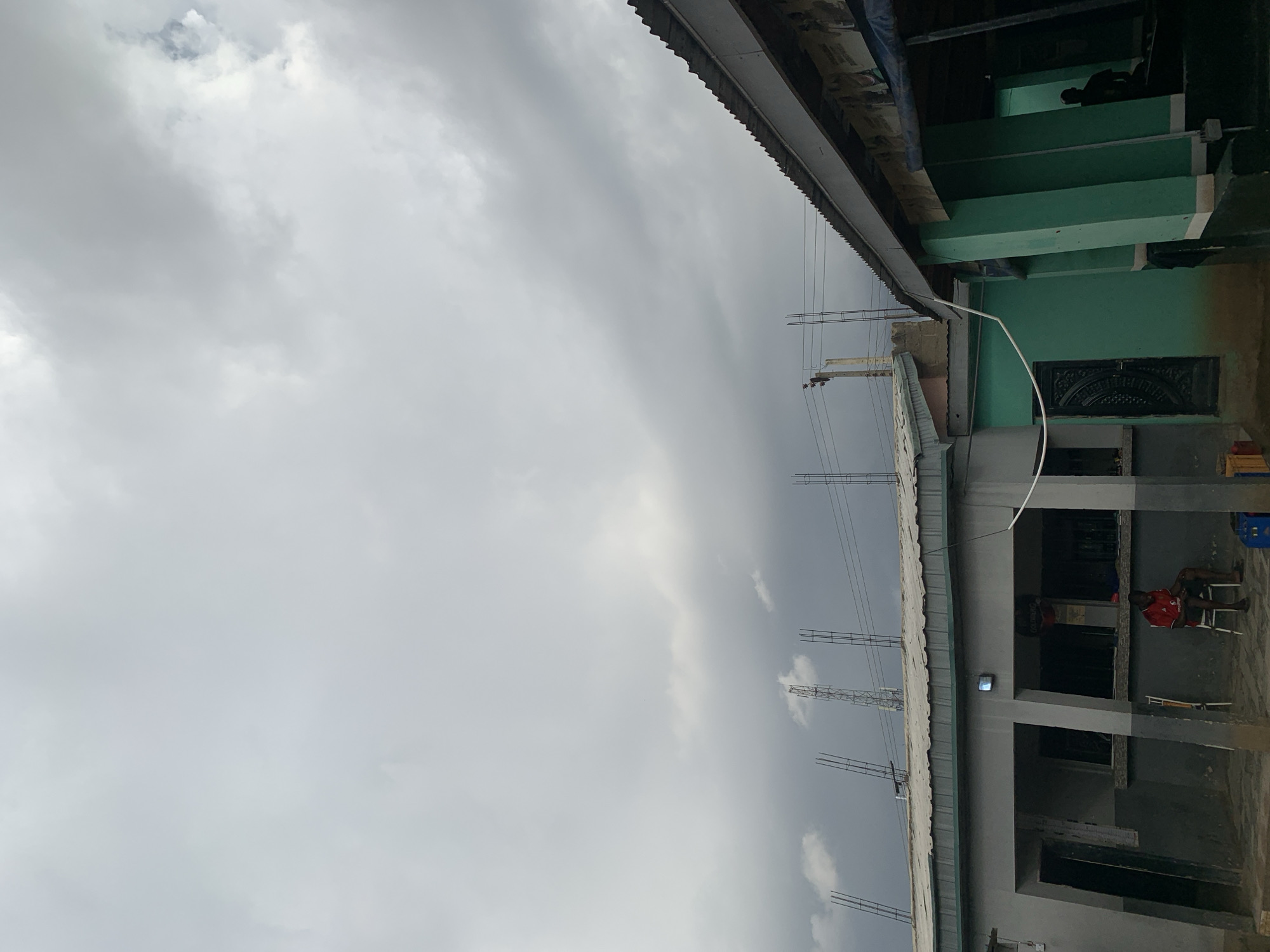 This image captures an outdoor scene under a heavily overcast, grey sky, suggesting a cloudy day. The perspective looks upwards towards several structures, indicating a possible construction site or an area with partially completed buildings.
On the left and extending through the center, electrical wires or communication lines are strung horizontally, supported by simple pole-like frameworks. Below these wires, a flat-roofed building, possibly under construction, is prominent. Its roof features multiple vertical metal rebars, hinting at further development. The building itself is made of concrete, with visible pillars and beams. A white pipe or conduit runs along its facade.
To the right, an adjacent building section is painted a distinctive teal or green color. It features a dark, ornate door with intricate patterns. Above this door, within the green structure, a dark silhouette of a person is visible, appearing to be seated or resting. At the very top right, a corrugated metal roof is partially visible, likely belonging to another nearby structure.
In the lower right portion of the image, within the grey building's framework, a person wearing a red shirt is seated on a white plastic chair. Another figure is faintly visible behind them.
The overall atmosphere is subdued due to the overcast sky. There is no clearly legible text visible within the image. Given the context of "New Stadium, Nigeria," these structures could be part of the stadium complex or surrounding ancillary buildings under development.