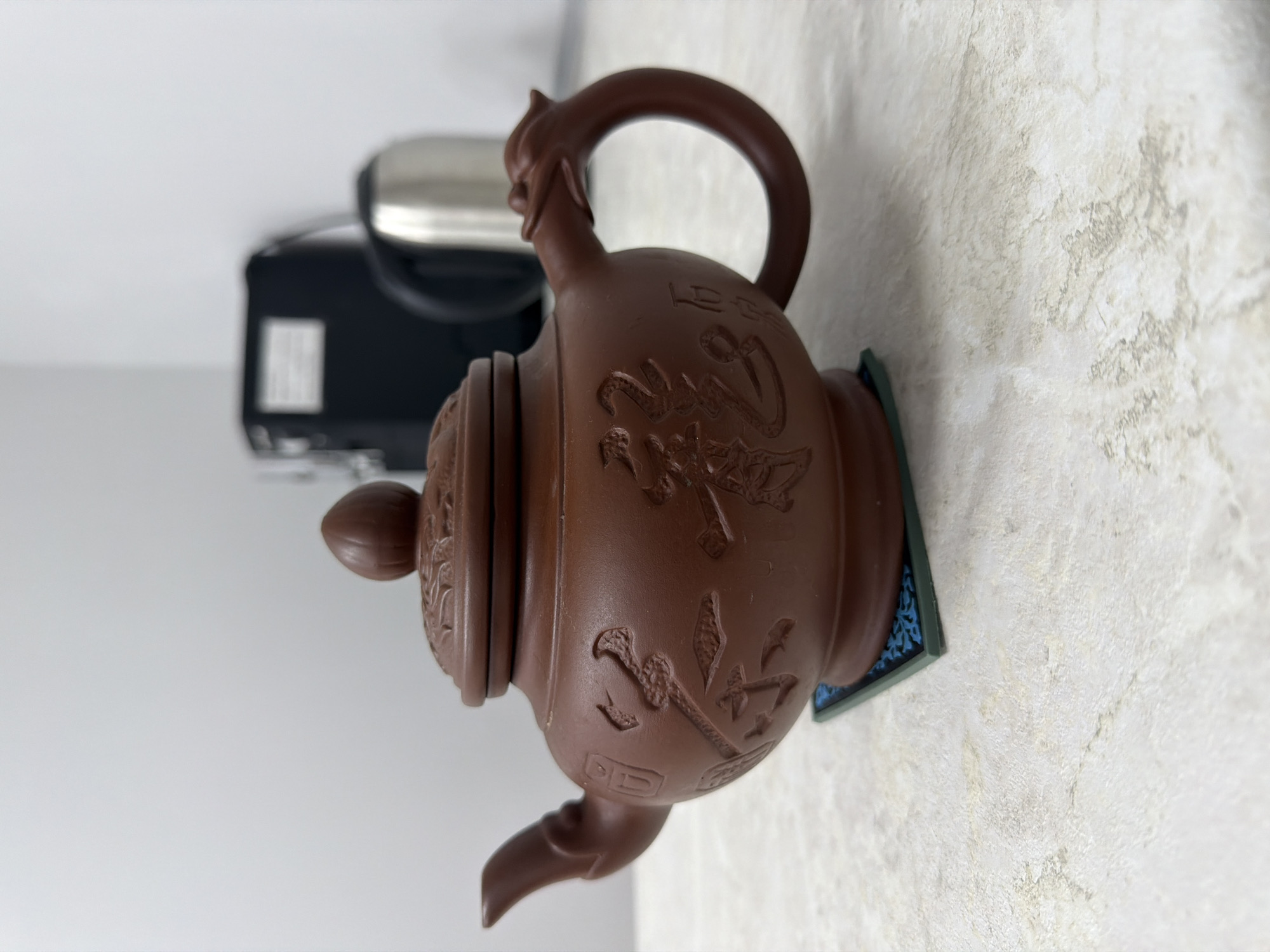 A brown teapot with Chinese inscriptions is sitting on a decorative blue and green tile coaster. In the background to the left is a small black appliance with a silver handle. The surface under the coaster is light-colored with a mottled pattern.
I cannot determine the location in Sarapul, Russia from this image.