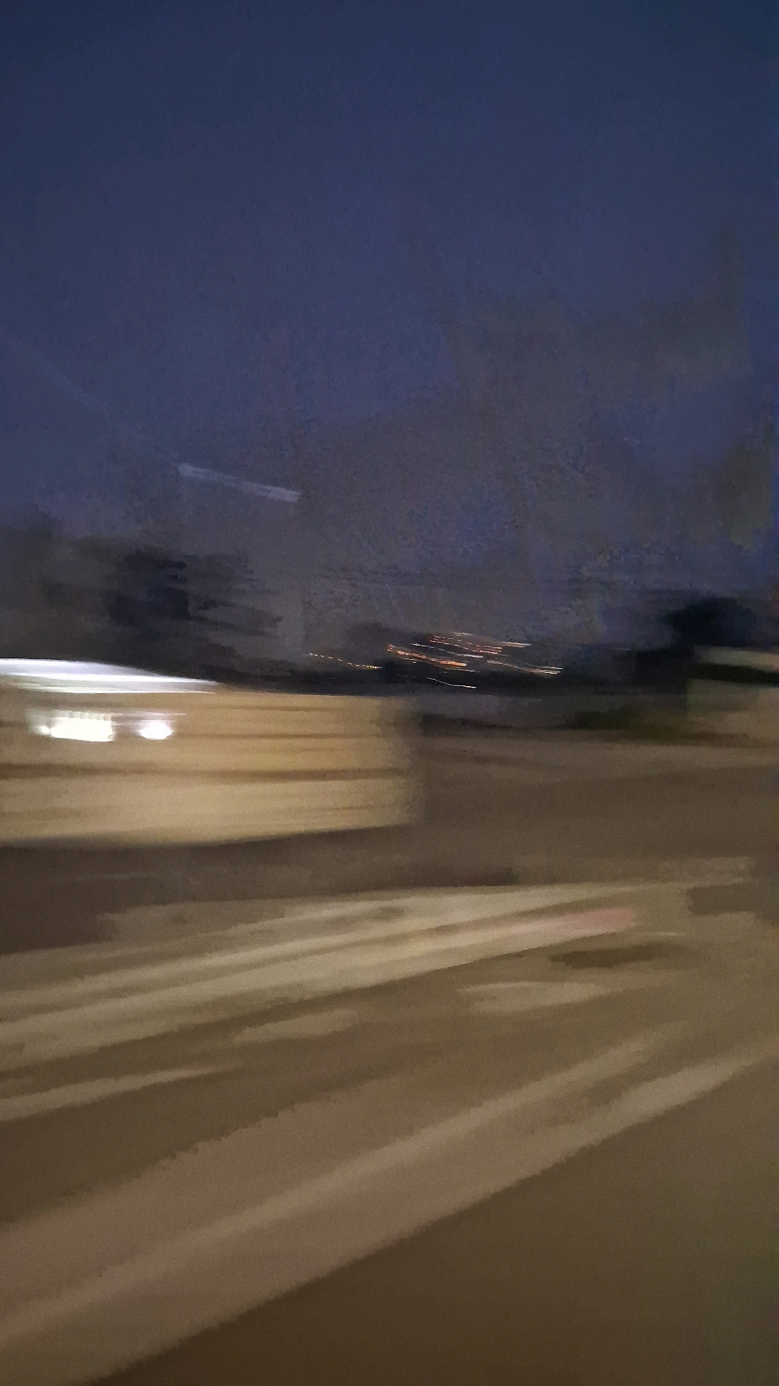 This image depicts a blurry, impressionistic view of a road at dusk or night. The sky is a deep, dark blue with hints of lighter clouds or haze. In the background, a blurred line of distant lights, likely from a city or town, stretches across the horizon. The foreground is dominated by a blurred representation of a road or a pedestrian crossing, with light-colored stripes running diagonally across it. To the left, there is a blurred, rectangular shape with bright lights, suggesting a building or vehicle. The overall impression is one of motion, as if the photograph was taken from a moving vehicle. The scene's location in Madurejo, Indonesia, is not visually discernible from the image itself, as the blurriness and generic urban/suburban elements could be found in many locations. There are no discernible people or specific interactions visible due to the motion blur and low light conditions.