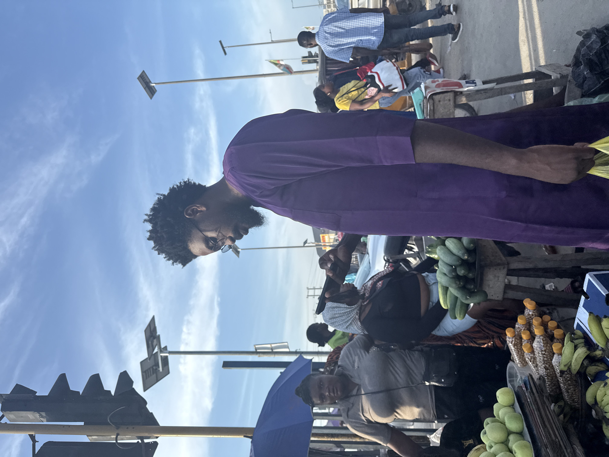 An outdoor street market or roadside vendor scene is depicted under a clear blue sky with sparse, wispy clouds. The lighting is bright and natural, consistent with either late morning or early afternoon, and the location is specified as Lekki, Nigeria.
In the immediate foreground, a dark-skinned man, approximately 25-35 years old, with an afro-textured hairstyle, a short beard, and eyeglasses, is visible from the chest up, positioned on the right side of the frame. He wears a purple, long-sleeved tunic. His gaze is directed downwards and slightly to his left. He holds a yellow plastic bag in his right hand, which hangs loosely by his side.
To the man's left, behind a market stall, a dark-skinned woman, also appearing to be a vendor, stands wearing a grey short-sleeved top and dark trousers. She has her hair pulled back and a black crossbody bag draped across her. Her gaze is directed forward and slightly to her right. To her immediate right, another woman, also positioned behind the stall, wears a black top and a patterned head covering. She holds a dark smartphone with both hands, appearing to interact with its screen.
The market stall in the mid-ground displays various goods. On a lower section, green apples are arranged on a tray, and clusters of yellow bananas are visible. Numerous small plastic bottles containing peanuts, each sealed with a yellow cap, are also displayed. A separate, taller wooden stand next to the woman in the head covering holds a pile of green cucumbers. A large blue umbrella is positioned on the far left, providing shade over part of the market area.
The background reveals typical urban street infrastructure. A black traffic light array is mounted on a yellow pole on the far left. Multiple tall poles, each topped with a solar panel and a streetlight fixture, extend into the sky. A national flag, consistent with the Nigerian flag given the specified location, is visible on a pole in the mid-background to the right of the main subject. Other pedestrians are present, including a woman in a yellow top carrying a large white and red bag, and a man in a blue and white checkered shirt facing away from the viewer. Partial views of vehicles and other indistinct urban structures are discernible further back. The ground surface appears to be paved, possibly asphalt or concrete.