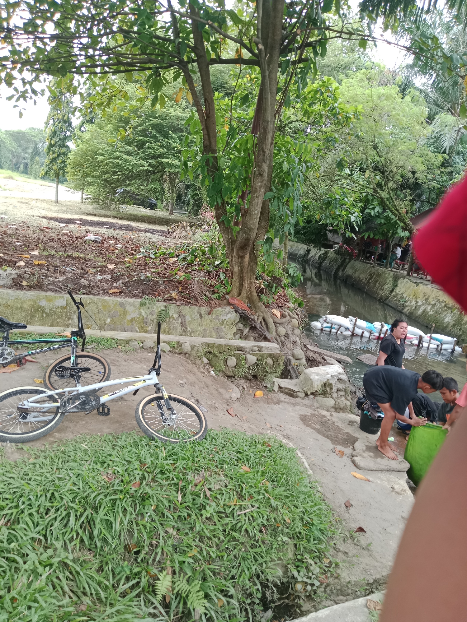 In the city of Binjai, Indonesia, a scenic outdoor setting features a small river or canal. Several individuals are present near the water's edge. One woman, wearing a black t-shirt, stands close to the canal. In the foreground, a young boy in a black t-shirt is bent over, seemingly interacting with a green bag. Another boy is partially visible beside him. Two BMX bicycles are parked on a dirt patch near a grassy area and a stone wall. The environment is lush with trees and vegetation, suggesting a park or recreational area. White sandbags are submerged in the canal, possibly for erosion control or to divert water. The presence of fallen leaves on the ground indicates an outdoor, natural environment.