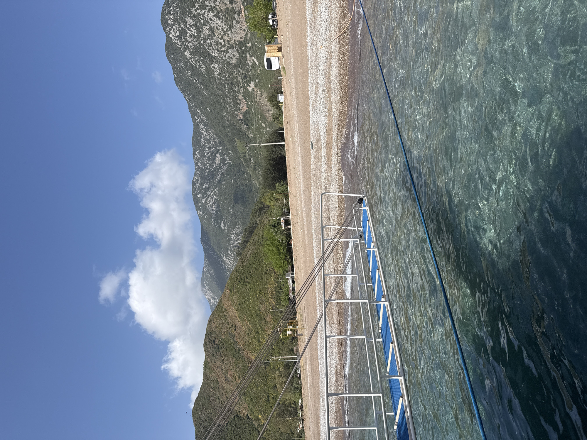 A landscape photograph taken at an angle shows a coastline in Kumluca, Turkey, with a rocky beach transitioning to clear water on the right. In the foreground, the blue and white railings of a boat or pier extend into the water. Along the beach, there are scattered vehicles and structures, possibly indicating a camping or recreational area.
Behind the beach, a mountain range rises, its slopes covered in greenery that transitions to exposed rock further up. A blue sky with scattered clouds is visible above the mountains. Power lines are also visible, stretching from the mountain area towards the beach.