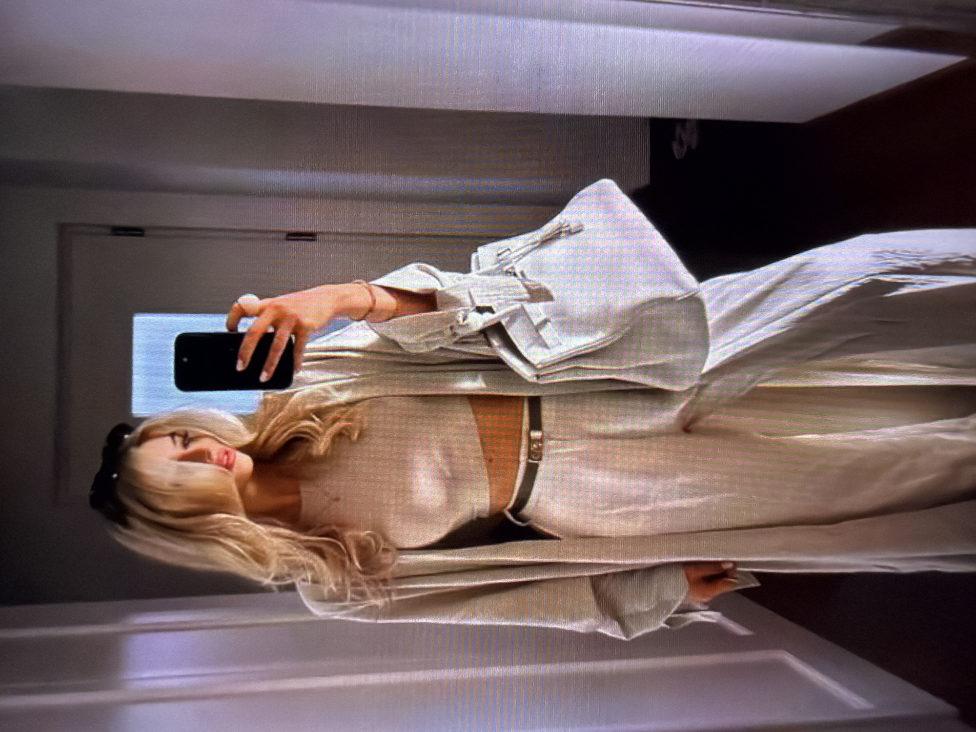 A young woman with blonde hair and light skin is taking a mirror selfie in what appears to be a room with a white door in the background. She is wearing a light-colored outfit consisting of a crop top and wide-leg pants, cinched at the waist with a dark belt. A light-colored blazer or jacket is draped over her shoulders. She is holding a black smartphone in her right hand, which is adorned with a large pearl ring. Her left hand is also visible, resting on the phone. The overall lighting suggests it is daytime. The room's simplicity and the presence of a door hint at an indoor setting, possibly a home or dressing room. The woman's expression is neutral, and the composition of the selfie focuses on her outfit and the presented pose.