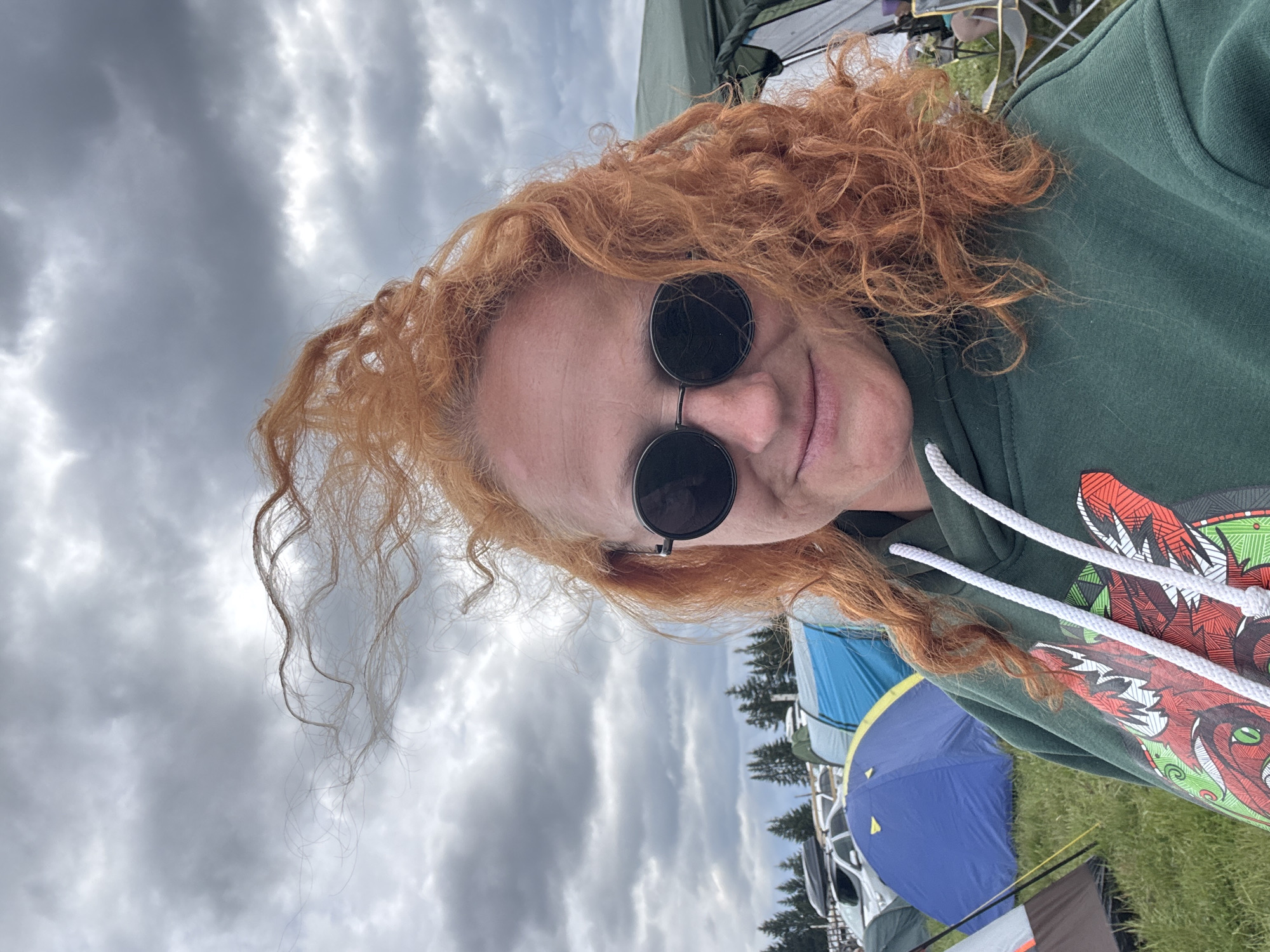 A close-up, tilted portrait features a woman with voluminous, curly, reddish-orange hair. She wears dark, round sunglasses and a dark green hooded sweatshirt with white drawstrings. A slight smile is visible on her lips. The front of her sweatshirt displays a vibrant, intricate graphic print, appearing to be a stylized creature or dragon in shades of red, white, and green.
The background depicts an outdoor setting, likely a camping area or festival grounds in Большая Кивара, Russia. The sky is overcast with a blanket of grey clouds, indicating a cloudy daytime. Various tents are visible, including a prominent blue dome tent, a blue and yellow tent, and a dark green tent, suggesting a multi-tent camp. Evergreen trees line the distant horizon, and parts of vehicles can be seen behind the tents. The ground is covered with green grass. The overall scene conveys a relaxed outdoor event. There is no visible text in the image.