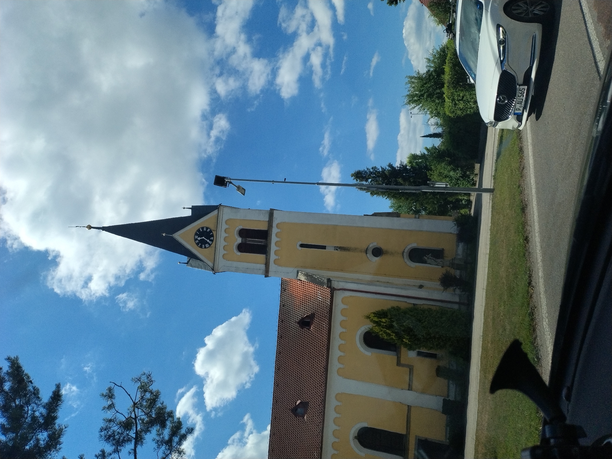This is a slightly angled, eye-level, outdoor photograph taken on a sunny day with scattered white clouds in a bright blue sky. The scene features a prominent yellow church with a reddish-brown tiled roof and a tall, dark steeple topped with a gold finial. The church has arched windows and decorative white trim. A clock face with Roman numerals is visible on the steeple. A tall street light stands to the right of the church. In the foreground, a white car with a license plate reading "P AU 565" is driving on a paved road. The car has a roof rack. To the left of the church, a dark silhouette of a car's interior is visible, including a black horn-like object and a windshield. Green grass and trees are present in the background and along the side of the road. The photograph appears to be taken in the town of Bližná, Czechia.