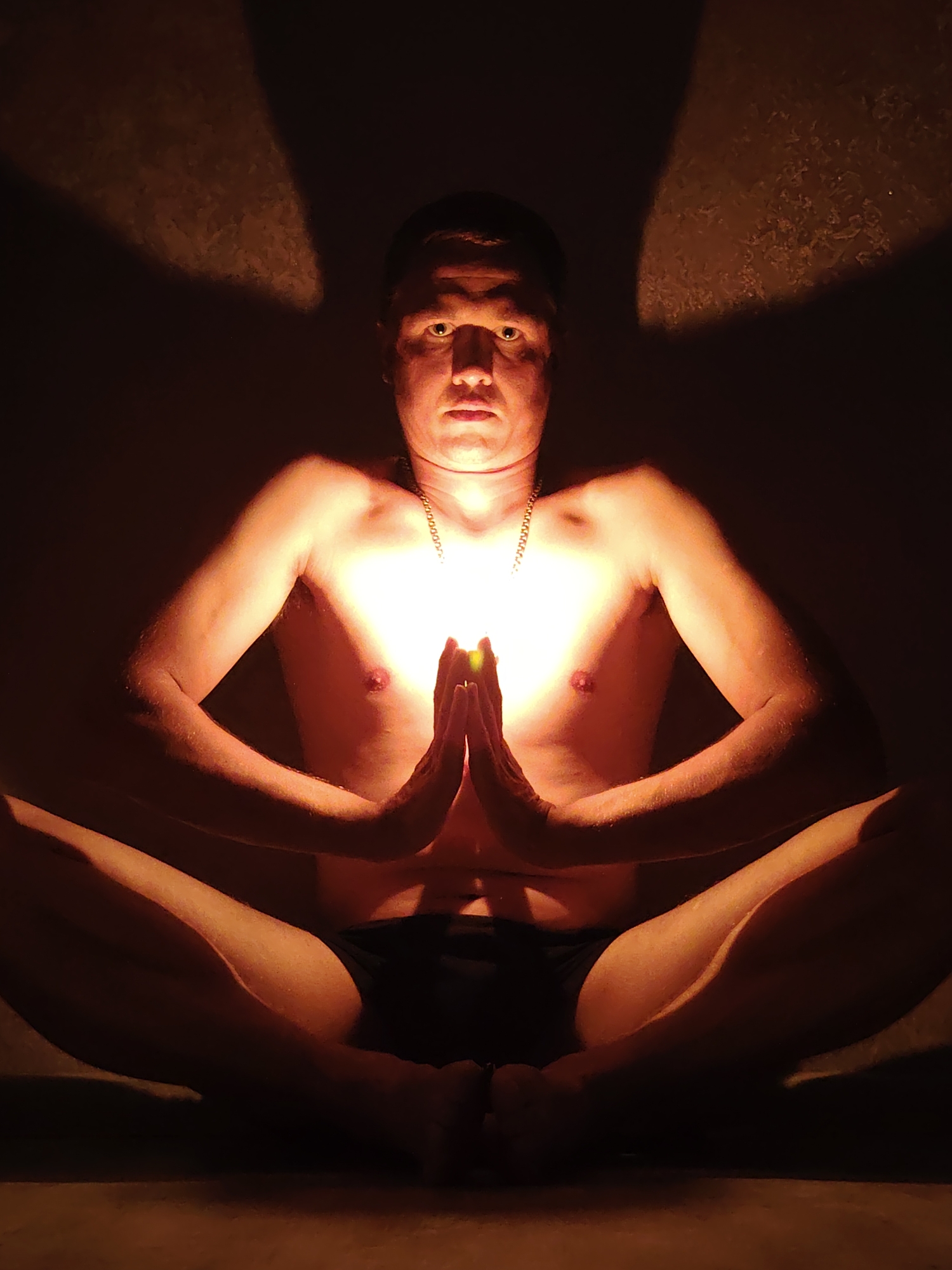 A person is seated in a lotus position in what appears to be an indoor setting. The person is bare-chested and wearing dark shorts. Their hands are pressed together in front of their chest, and a bright light emanates from between their palms, illuminating their torso and face. The background is dark, with a textured wall on which shadows are cast. The image suggests a moment of meditation or spiritual practice, possibly in a dimly lit room in Chaikovsky, Russia.
