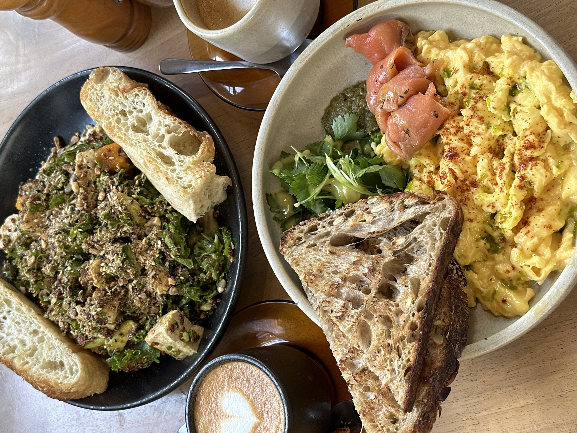 A top-down view on a light wooden surface reveals a breakfast or brunch scene in Unknown, Unknown. On the left, a dark plate holds a vibrant salad featuring green leafy vegetables, diced tofu, and what appears to be roasted butternut squash. The salad is topped with a sprinkle of seeds and paired with a slice of white bread.
Adjacent to the salad, a small cup of latte displays a heart-shaped foam design. To the right, a second bowl contains scrambled eggs speckled with green herbs, accompanied by several slices of smoked salmon, a dollop of pesto, and a sprig of fresh herbs. The bowl is served with a slice of rustic, crusty bread.
In the upper part of the image, out-of-focus items suggest condiments, possibly salt and pepper mills, and another cup of coffee. The setting suggests a cafe or restaurant environment.
