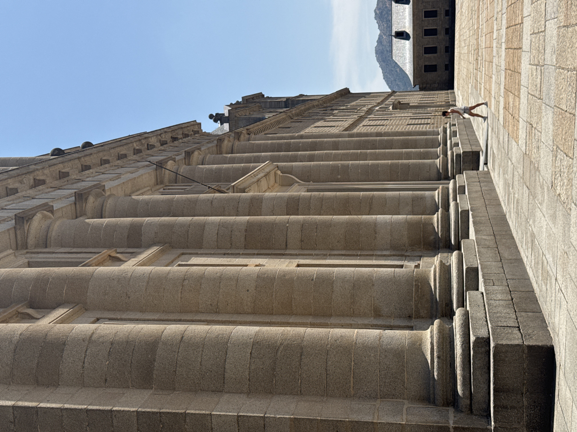 This image, taken from a low angle and rotated 90 degrees counter-clockwise, shows a person standing on the large, stone steps of a grand building. The building appears to be made of granite blocks and features classical architectural elements like columns and ornate cornices. In the background, a mountain range is visible, along with more of the building which has a dark, monolithic appearance with visible windows. The sky is clear and blue, suggesting a sunny day. The person, dressed in light-colored shorts and a white t-shirt, is leaning forward with one leg extended, as if in mid-stride or posing. The overall impression is one of scale and grandeur, with the human figure dwarfed by the immense architecture. The location is identified as San Lorenzo De El Escorial, Spain. The time of day is likely midday or early afternoon due to the bright sunlight and strong shadows. There is no visible text in the image.