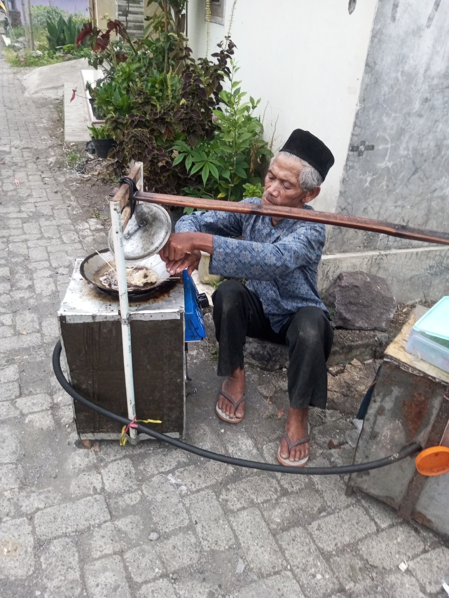 An elderly man with graying hair and a black peci cap is preparing food outdoors, presumably in the city of Andongsili, Indonesia. He is seated on the ground, wearing a patterned blue shirt and dark pants, with flip-flops on his feet. In front of him is a portable cooking setup. A metal box, likely a stove or cooler, supports a round frying pan where food is being cooked. The man is actively engaged in cooking, holding utensils and manipulating skewers of food in the pan. A large metal lid is partially open above the pan. A black hose, possibly for gas, is connected to the cooking apparatus and secured with yellow and red ties to a white pole. The ground is paved with interlocking gray bricks, and there is a gray wall and lush green foliage in the background. Rocks and other metallic containers are also visible around the cooking area.