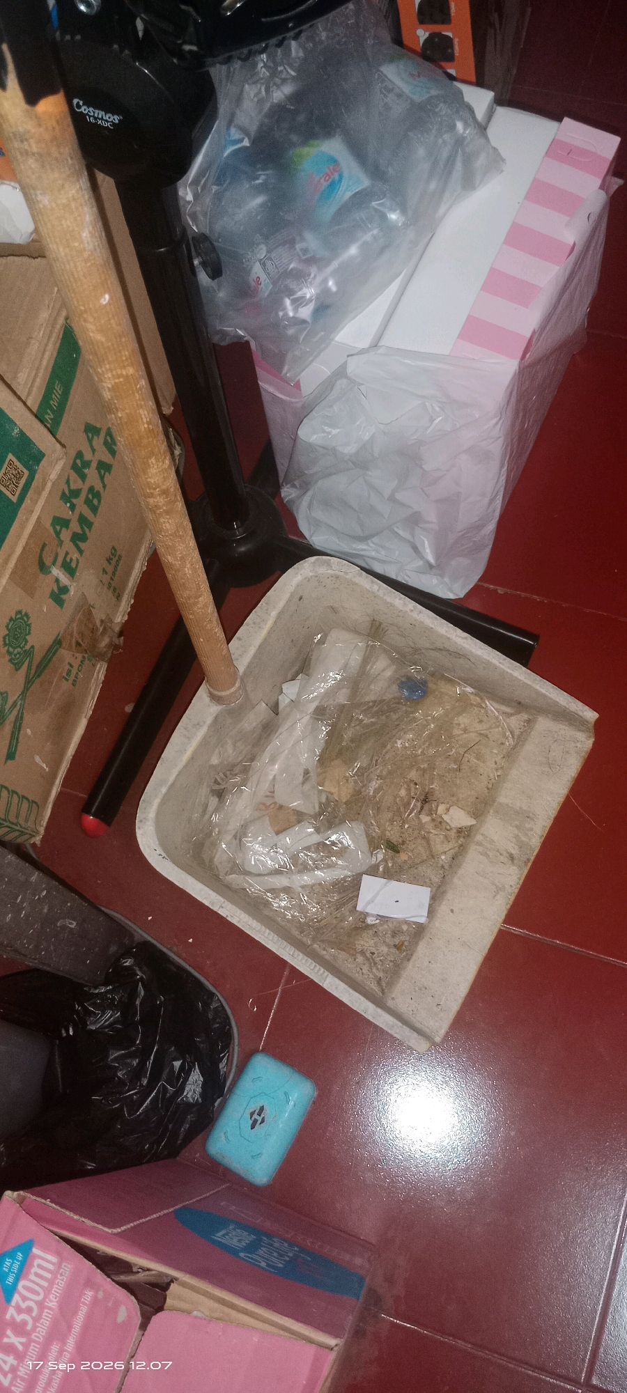The image depicts a close-up, high-angle view of a dusty dustpan filled with debris, including dirt, small pieces of paper, and other unidentifiable waste. The dustpan is made of a light-colored material, possibly plastic or metal, and has a wooden handle attached to a black stand. To the left of the dustpan, a portion of a cardboard box with green lettering that reads "CAKRA KEMBAR" is visible. Behind the dustpan, several plastic-wrapped items are stacked, with one item featuring pink and white stripes. In the lower left quadrant, a black trash bag and a bright blue rectangular object, possibly a soap or a small container, are present on a red tiled floor. The overall scene appears to be indoors, potentially in a storage area or a less-maintained part of a dwelling. The timestamp "17 Sep 2026 12:07" is visible in the bottom left corner, suggesting the image was taken on that date and time. Without further information, it is impossible to definitively place this scene within the city of Klaten, Indonesia, or provide any specific details regarding people, actions, or interactions.