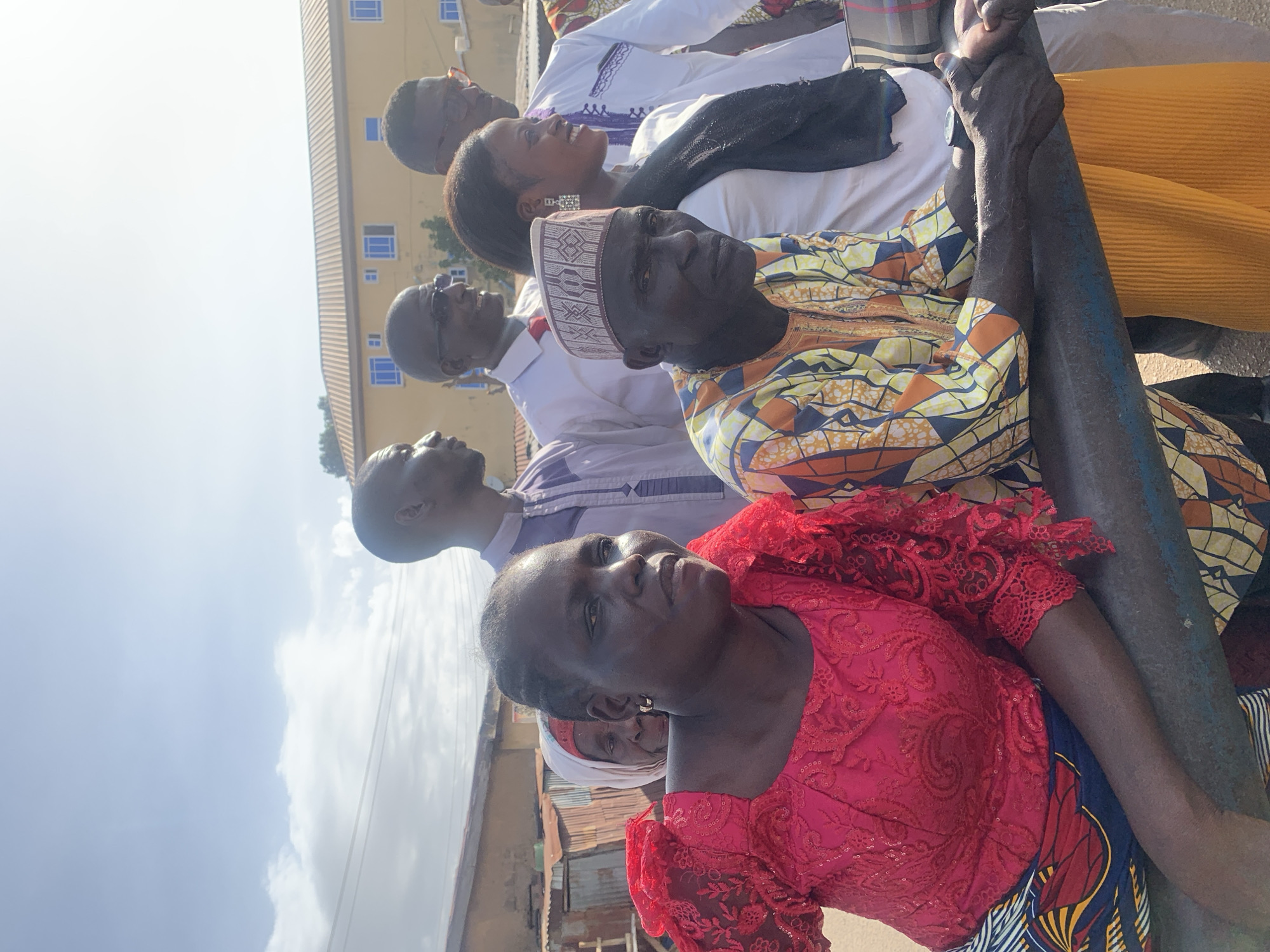 A low-angle shot captures a diverse group of people, predominantly adults of African descent, gathered outdoors in what appears to be Jos, Nigeria. The image is bathed in bright daylight, with a clear to partly cloudy sky visible in the upper left.
In the immediate foreground, to the left, a woman is seen from the chest up, wearing a vibrant red lace top with intricate patterns and a colorful patterned skirt. She looks slightly upwards and to her right, her expression appearing attentive. Behind her, partially obscured, another woman in a white headscarf is visible.
To the woman's right, and slightly behind her, a man with dark skin wears a traditional attire featuring a patterned yellow, orange, brown, and blue top and a distinctive white and brown embroidered cap. He looks directly forward with a serious expression. His right arm, extended forward, is visible.
Further back in the group, several other individuals are looking upwards or intently forward. A man in a white shirt and dark glasses is seen looking up and to his left. Behind him, a woman with dark hair and large, dangling earrings is smiling and looking upwards, wearing a white top and a black shawl. Another man, wearing white traditional attire with purple embroidery and dark glasses, is also visible further back, looking upwards.
On the far right, a sturdy, dark-colored pole or pillar stands vertically, partially obscuring some of the background. In the background, a light-colored building with multiple blue-paned windows suggests an urban or semi-urban setting. Further left in the background, hints of corrugated metal roofs and other structures are visible, characteristic of local architecture.
The group's collective upward gaze suggests they are observing an event, listening to a speaker, or looking at something of interest above them. No visible text is present in the image.