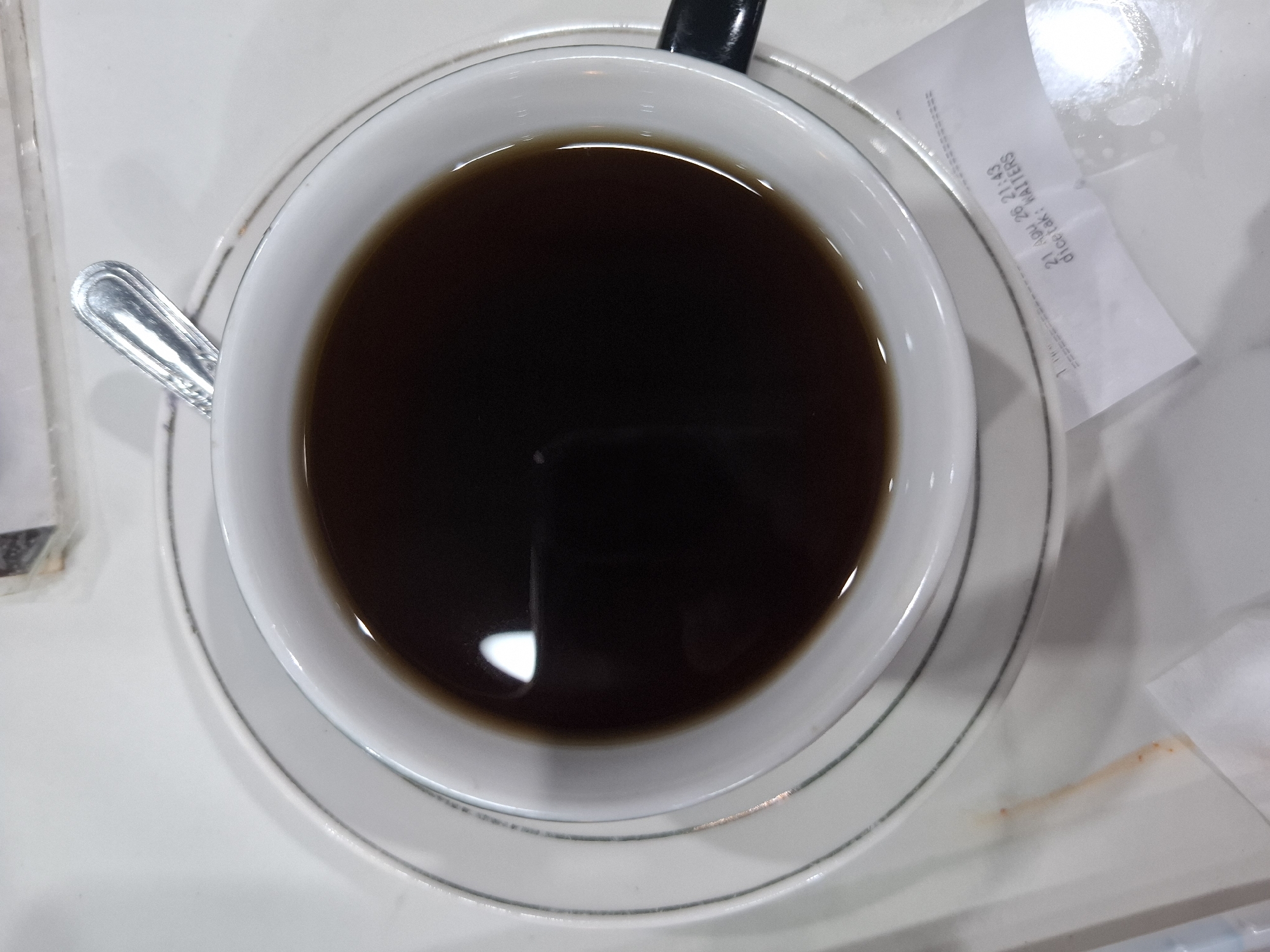 A top-down, medium shot captures a close-up of a white ceramic cup filled with dark coffee, resting on a matching saucer. A silver spoon is placed diagonally across the rim of the cup, with its handle extending beyond the cup. The coffee surface reflects a bright light source. A white receipt with printed text is partially visible beneath the saucer. The background consists of a white, textured surface, suggesting a table. The receipt displays a date and time stamp: "21 Agu 26 21:43" and "dicetak: WAITERS". The scene is devoid of any human presence, activity, or clear indication of its specific location within Kampongbaro, Indonesia, other than what might be inferred from the receipt's text, which is not a definitive geographical locator.