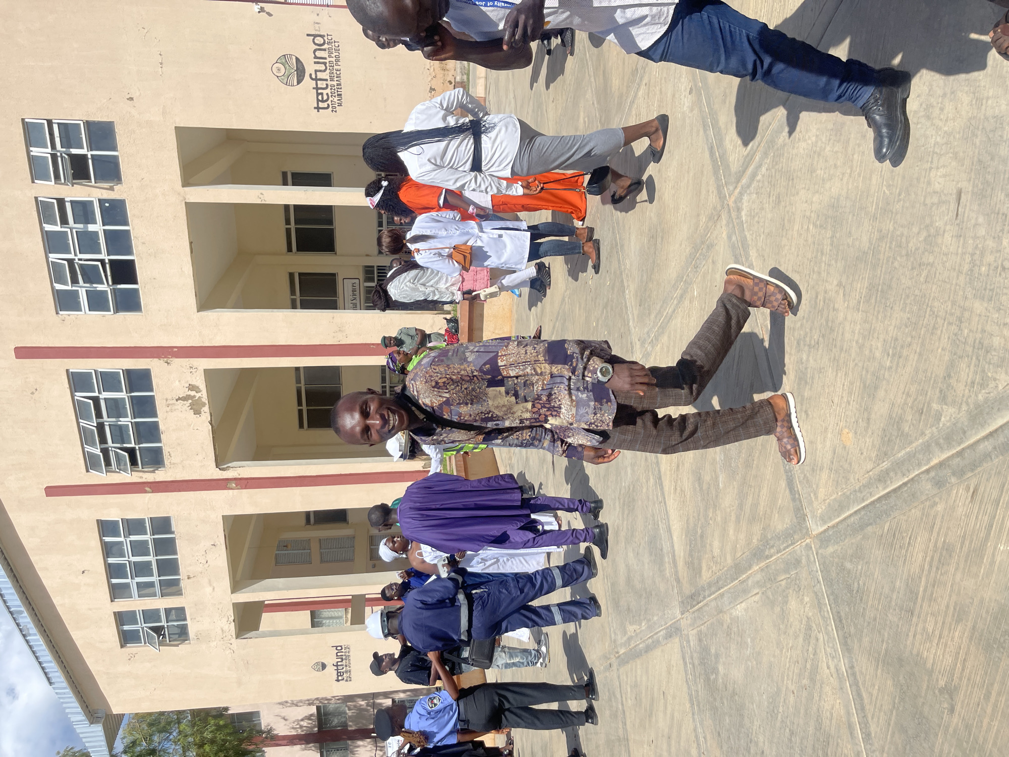 This is a medium shot, taken outdoors, of a group of people in front of a beige-colored, multi-story building. The building has a series of windows on its upper floors and a large, open-air structure with columns on the ground level. A sign on the building reads "tetfund 2017-2020 MERGED PROJECT MAINTENANCE PROJECT." The ground is paved with large concrete slabs.
In the foreground, a man with dark skin is smiling and looking at the camera. He is wearing a dark, patterned shirt and plaid pants, and is walking on brown and white sandals. Behind him, other people are visible. To the left, a person in a police uniform is talking to another individual. In the center background, several people in traditional attire are gathered, including one person in a purple robe and another in white. To the right, a person wearing a white shirt and grey skirt is walking away from the camera. The lighting suggests it is a sunny day, casting shadows on the ground. The scene appears to be at a university or public institution in Rafin Par, Nigeria, given the presence of the TETFUND sign, a Nigerian federal government agency.