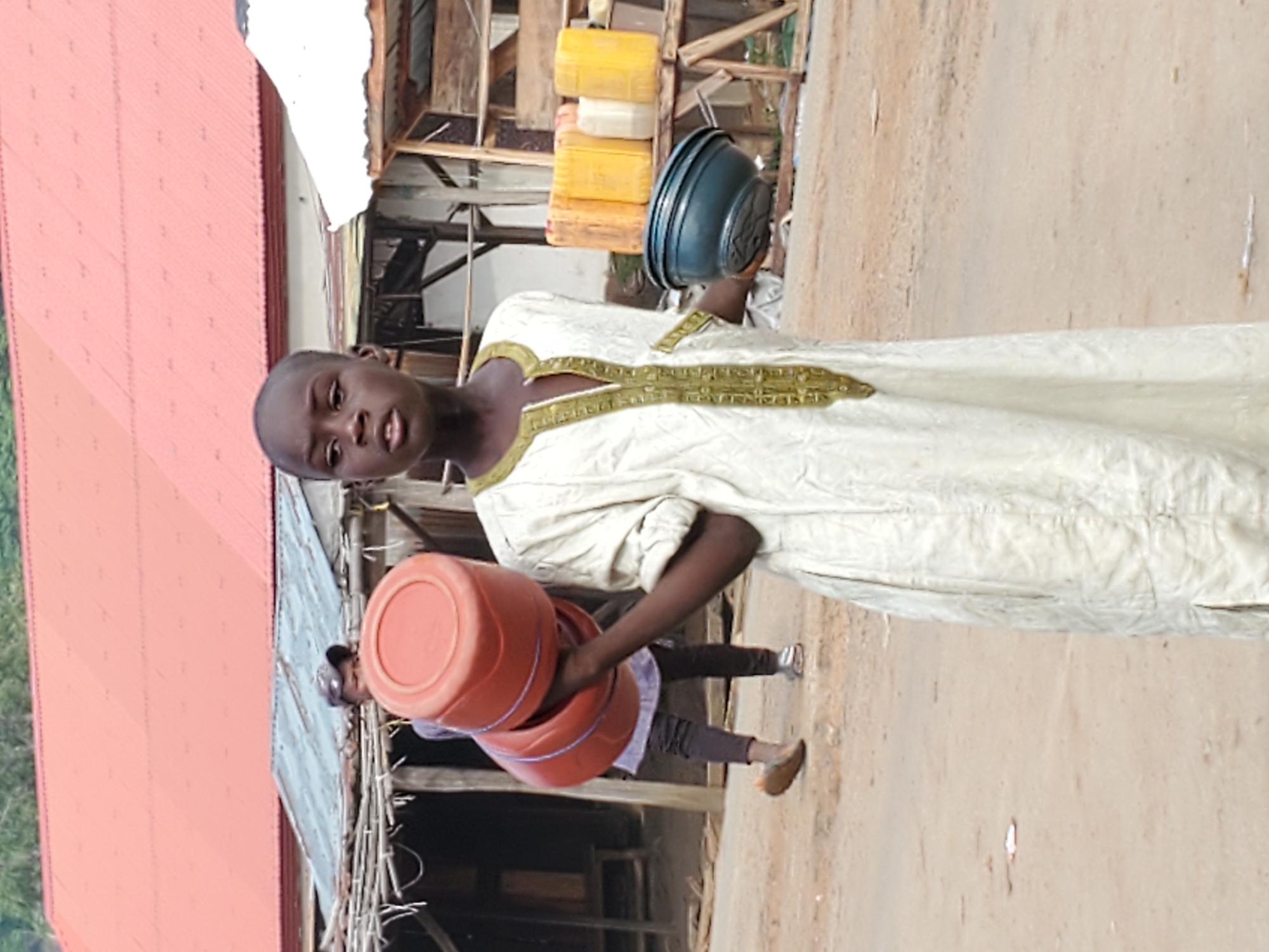In Mararaba, Nigeria, a young person stands on a dirt road next to a building with a red roof. They are carrying two red buckets in one arm and a dark blue bowl in the other. The person is wearing a long white garment with gold trim around the neckline. Another person is walking in the background. In the background there is also a wooden structure and yellow jugs.