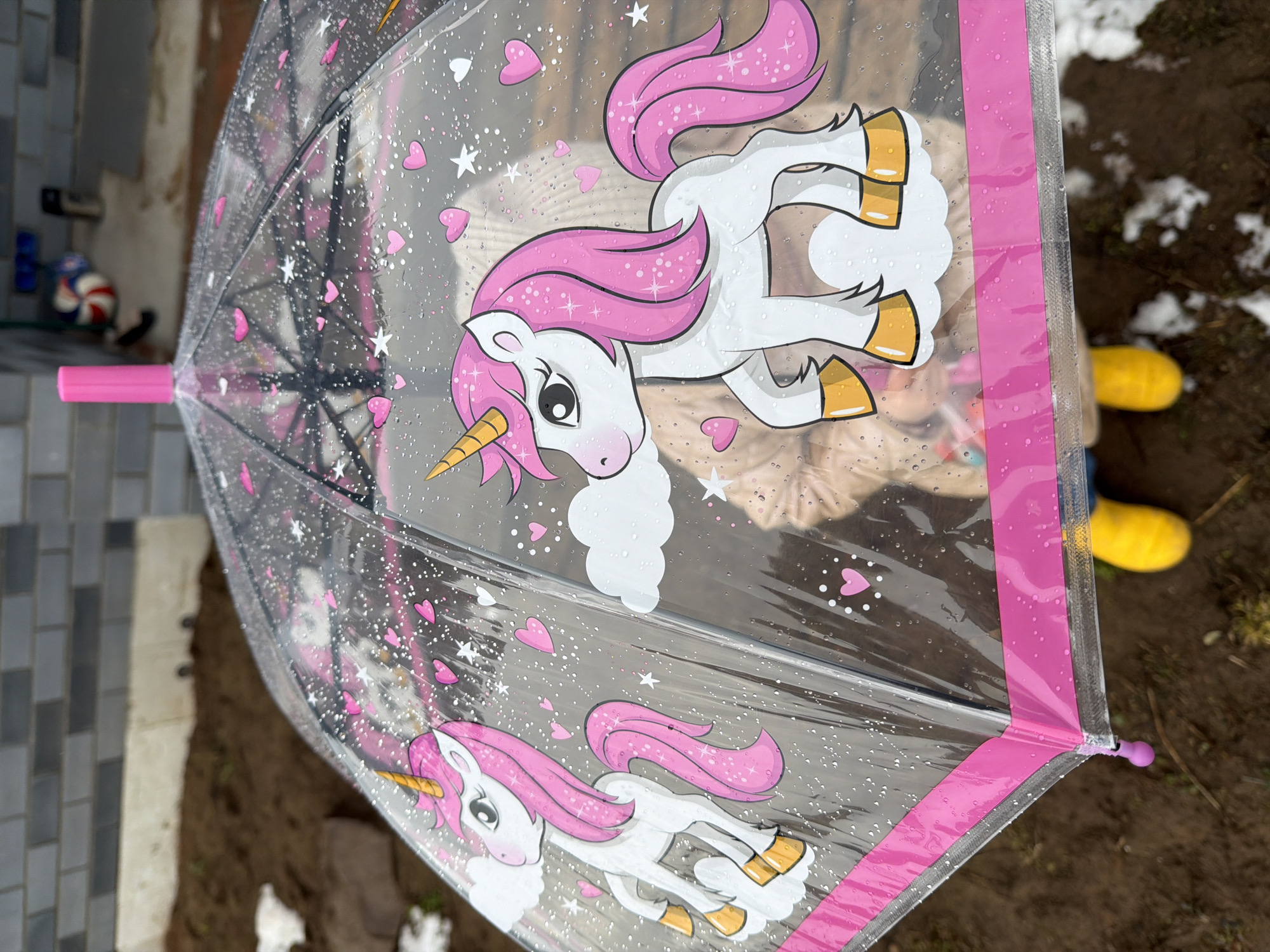 A clear, dome-shaped umbrella with a pink handle and trim sits outdoors on a partly cloudy day, reflecting the image of a child wearing a tan jacket and yellow boots. The umbrella is decorated with images of white unicorns with pink manes, tails, and golden hooves, along with pink hearts and white stars. The ground beneath the umbrella appears to be dirt and grass, with patches of melting snow visible. In the background, there are glimpses of a brick wall and a few toys. The photograph was taken in the city of Chaikovsky, Russia.