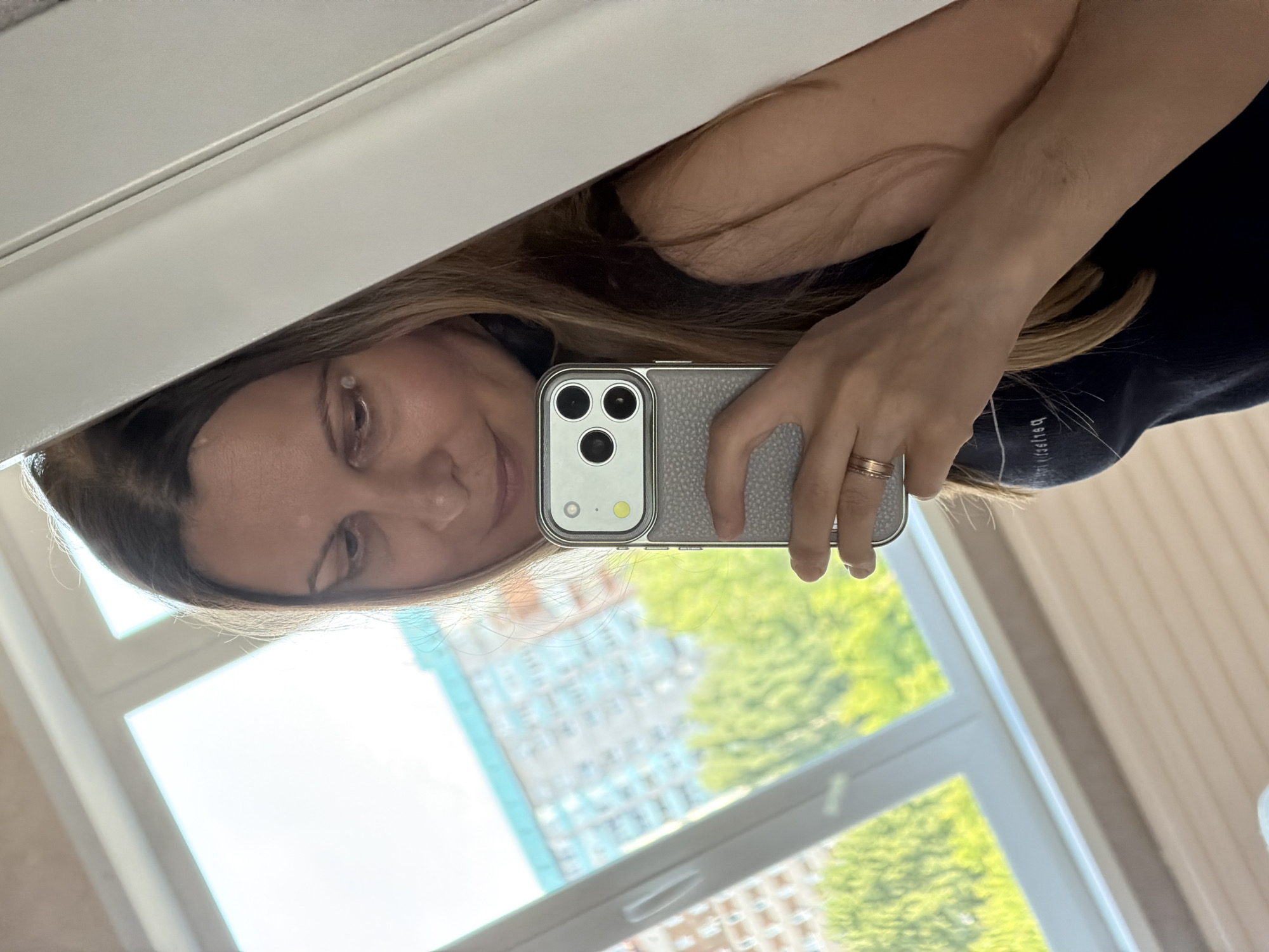 A woman is taking a selfie in a mirror. She is holding a silver smartphone in her right hand. She is wearing a dark blue top and a ring on her left ring finger. Her hair is long and brown. She is standing in front of a window that looks out onto a city scene with buildings and trees. The room appears to be an apartment in Izhevsk, Russia, judging by the architectural style of the buildings visible through the window.