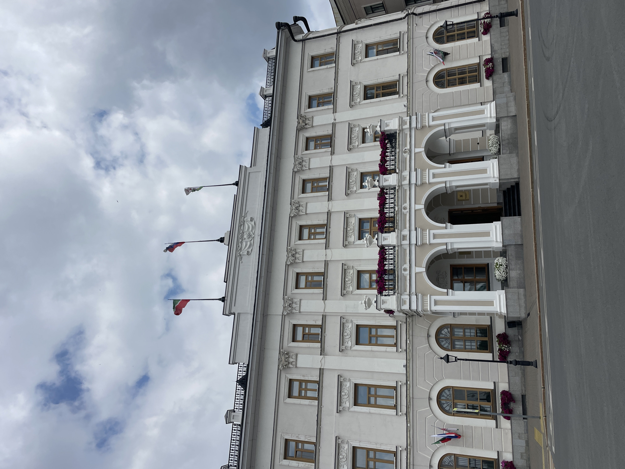 The image depicts a grand, light-colored building with a neoclassical architectural style, likely a government or administrative building, situated in the city of Kazan, Russia. Three national flags are prominently displayed on flagpoles atop the building, indicating a significant official location. The building features a white facade with numerous windows, many of which are arched. A balcony, adorned with floral arrangements, extends from the central section of the upper floor. The ground floor has a covered entrance with arched columns. The street in front of the building is paved, with a sidewalk and a lamppost. The sky is overcast with clouds, suggesting it is not a perfectly sunny day. The overall scene conveys a sense of civic importance and historical architecture.