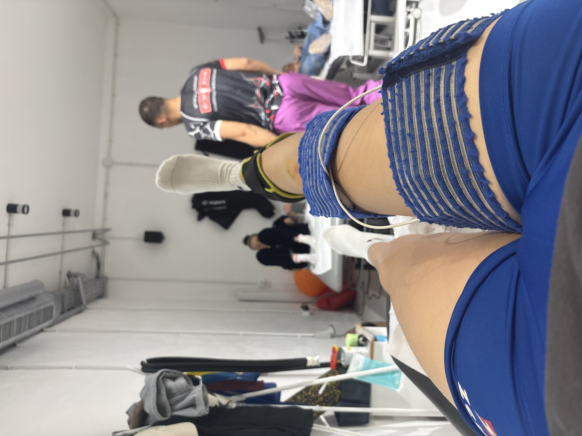 The image shows an interior scene, likely in a physical therapy or sports rehabilitation setting. In the foreground, a person wearing blue athletic shorts and a compression wrap on their thigh is visible. The wrap is light blue and white.
In the midground, there are two other people: one wearing a black and red athletic shirt and lilac pants and another person seated and wearing black clothes. An orange exercise ball can be seen near them.
In the background, medical equipment and other miscellaneous items are visible. The overall environment appears to be a clinical or rehabilitation space with white walls. The image was taken indoors, but there is no specific information to confirm the location in Sofia, Bulgaria.