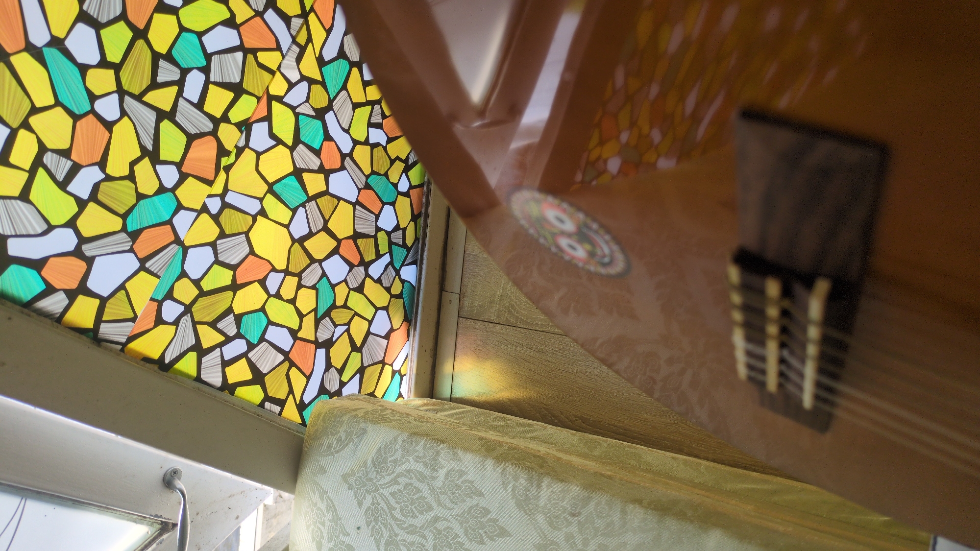 The image shows a view looking up from a surface covered with a patterned cream-colored fabric. To the left is a window with a geometric stained glass design in shades of yellow, orange, green, and white, separated by black lines. The window frame is visible. Reflected in the brown, varnished surface above is a portion of the stained glass window. A sticker or emblem and a part of a stringed instrument, possibly a guitar or lute, including its bridge and strings, are also reflected in the varnished surface. A strip of rainbow light is present on the brown surface between the window and reflected instrument. The location is Da Lat, Vietnam.